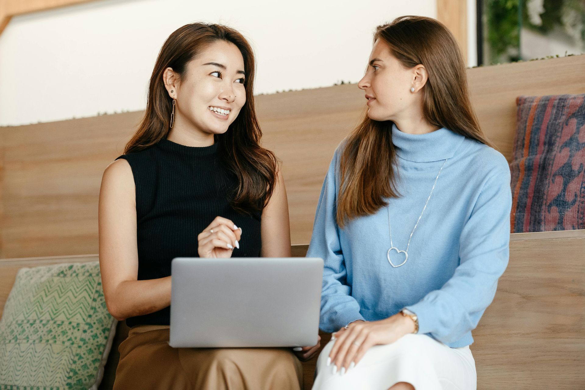 Two people sit side-by-side on a wooden bench, one pointing at a laptop screen while the other looks on and listens.