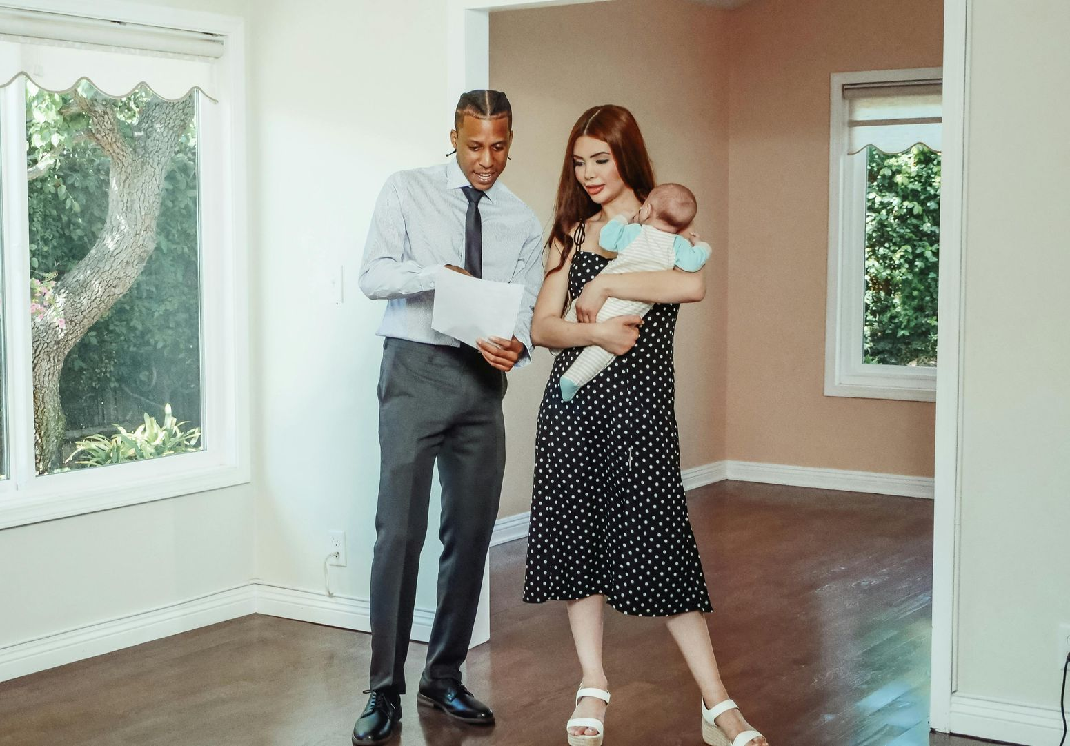 A person in a shirt and tie shows papers to a person holding an infant in a bright, empty room with hardwood floors.