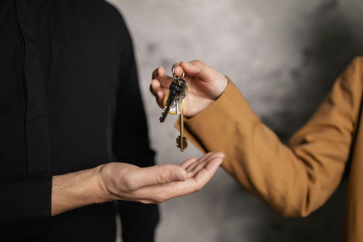 One person in a tan jacket hands a set of keys to another person in a black shirt against a textured gray background.