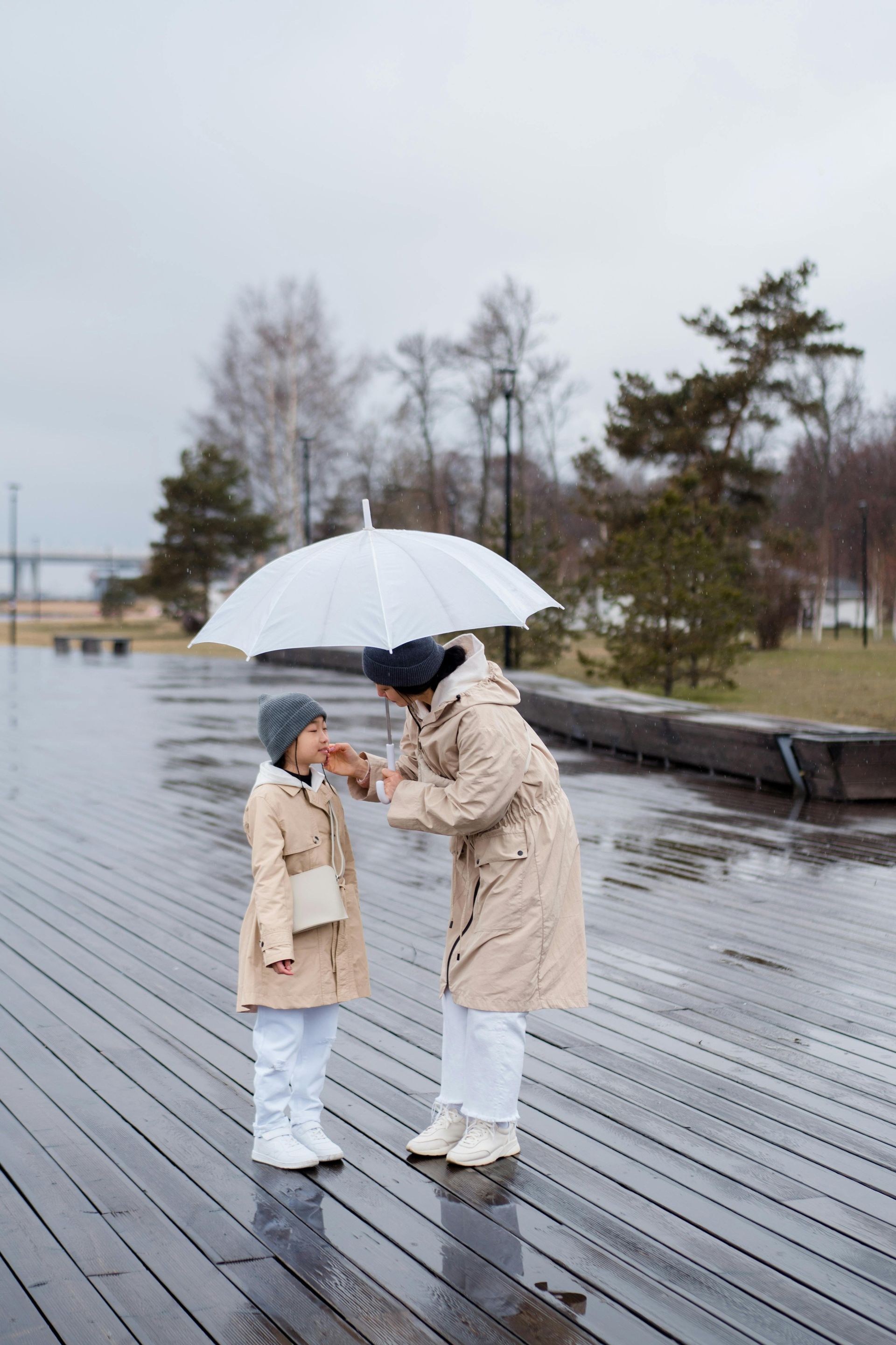A person leans down to tuck a scarf under a child's chin while standing under a white umbrella on a wet wooden pier.
