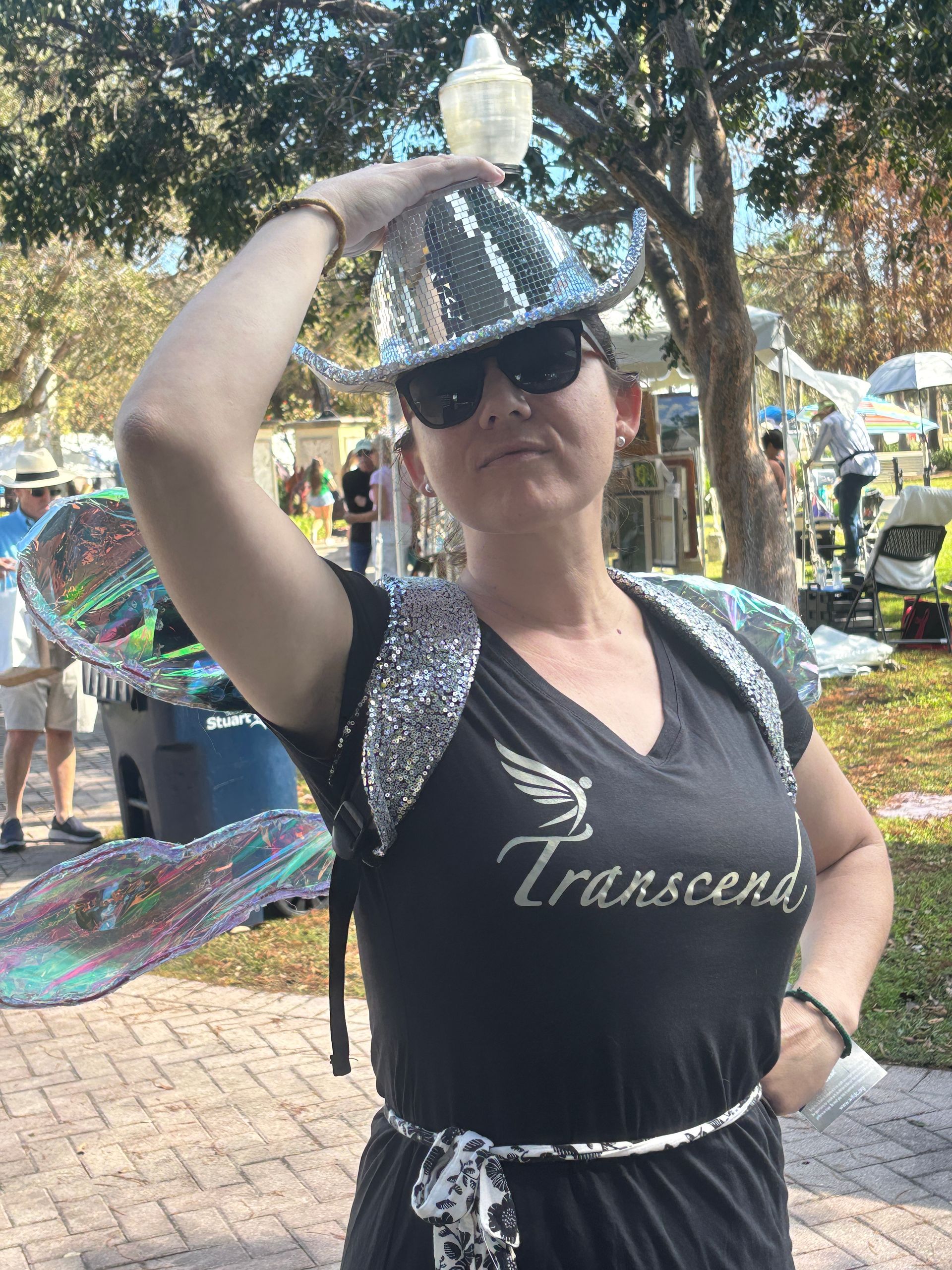 Person in sunglasses posing outdoors in a black sequined shirt under trees at a festival