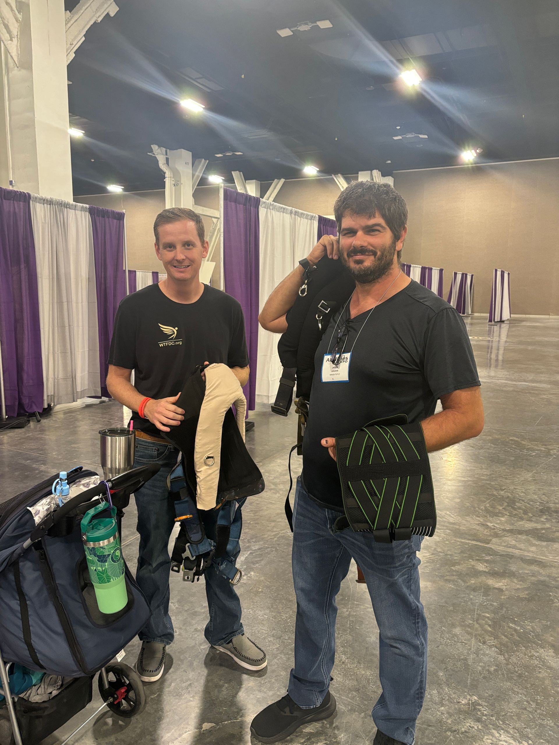 Two people standing in a convention hall with coat racks and a stroller, one holding a drink and the other smiling.
