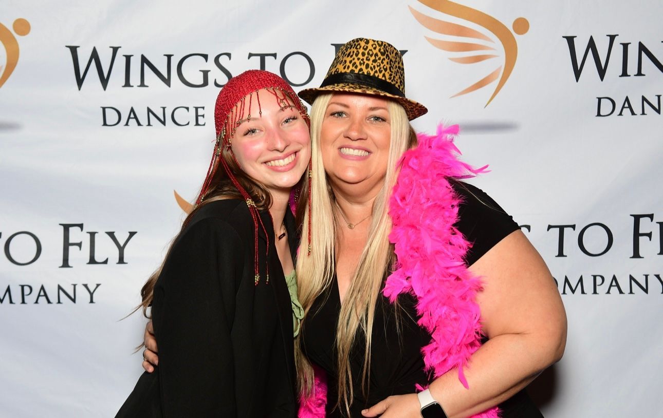 Two smiling people pose in front of a Wings to Dance backdrop, wearing black outfits and bright feather boas.
