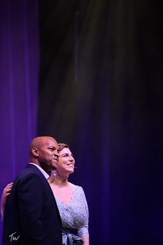 Two people in formal attire standing onstage under purple lighting, smiling toward the audience