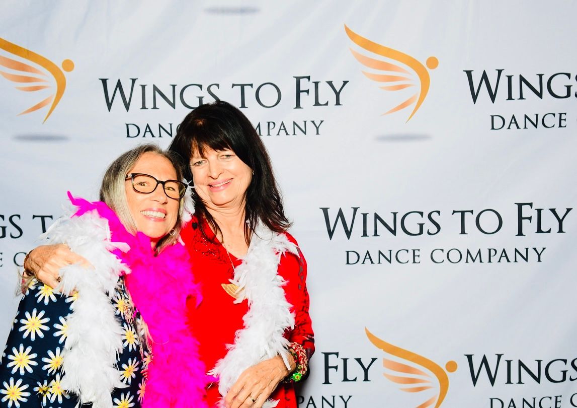 Two people smiling at a Wings to Fly Dance Company event backdrop, wearing bright feather boas
