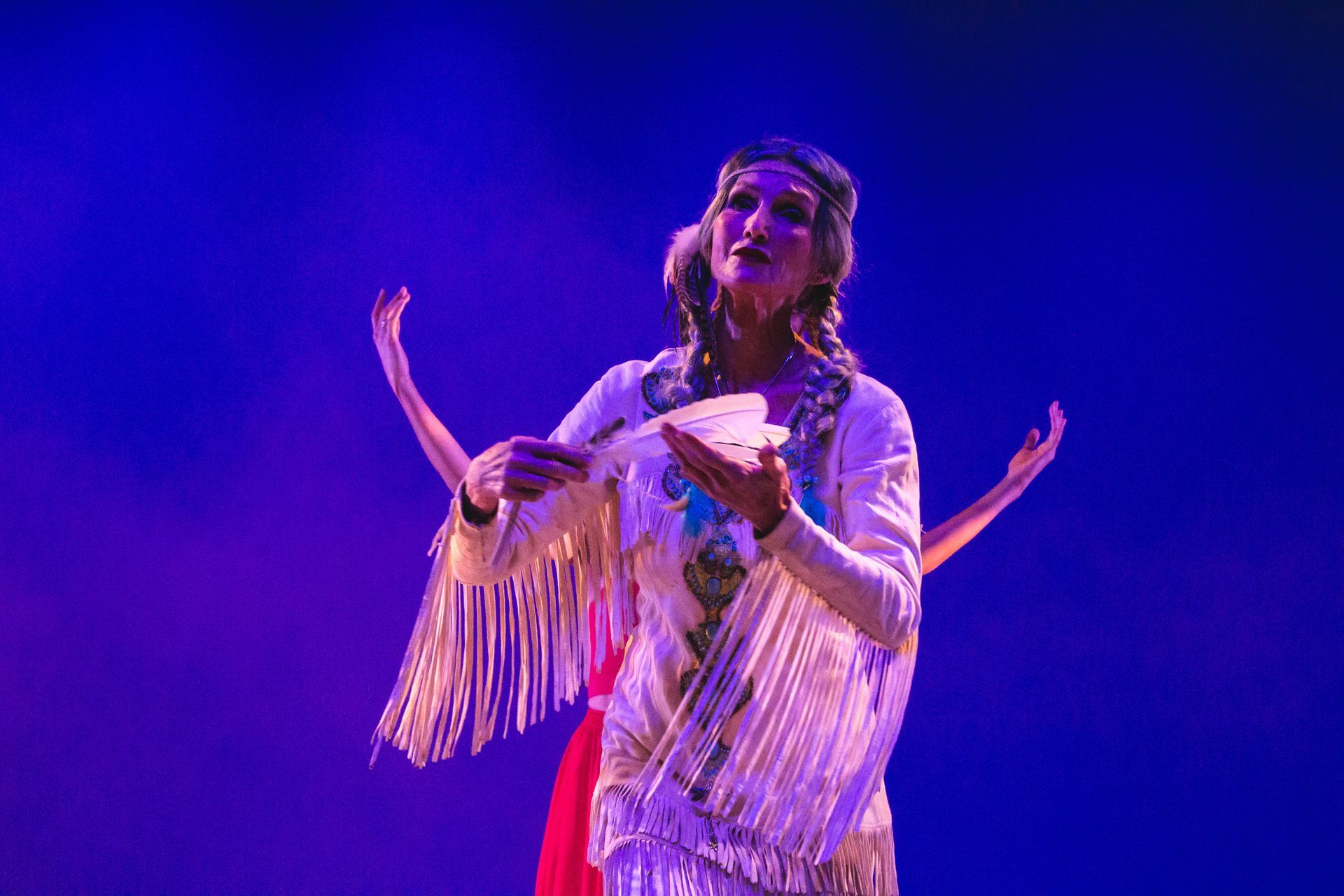 Woman in white fringed shawl, holding a fan, onstage with arms raised, lit by blue and purple lights.