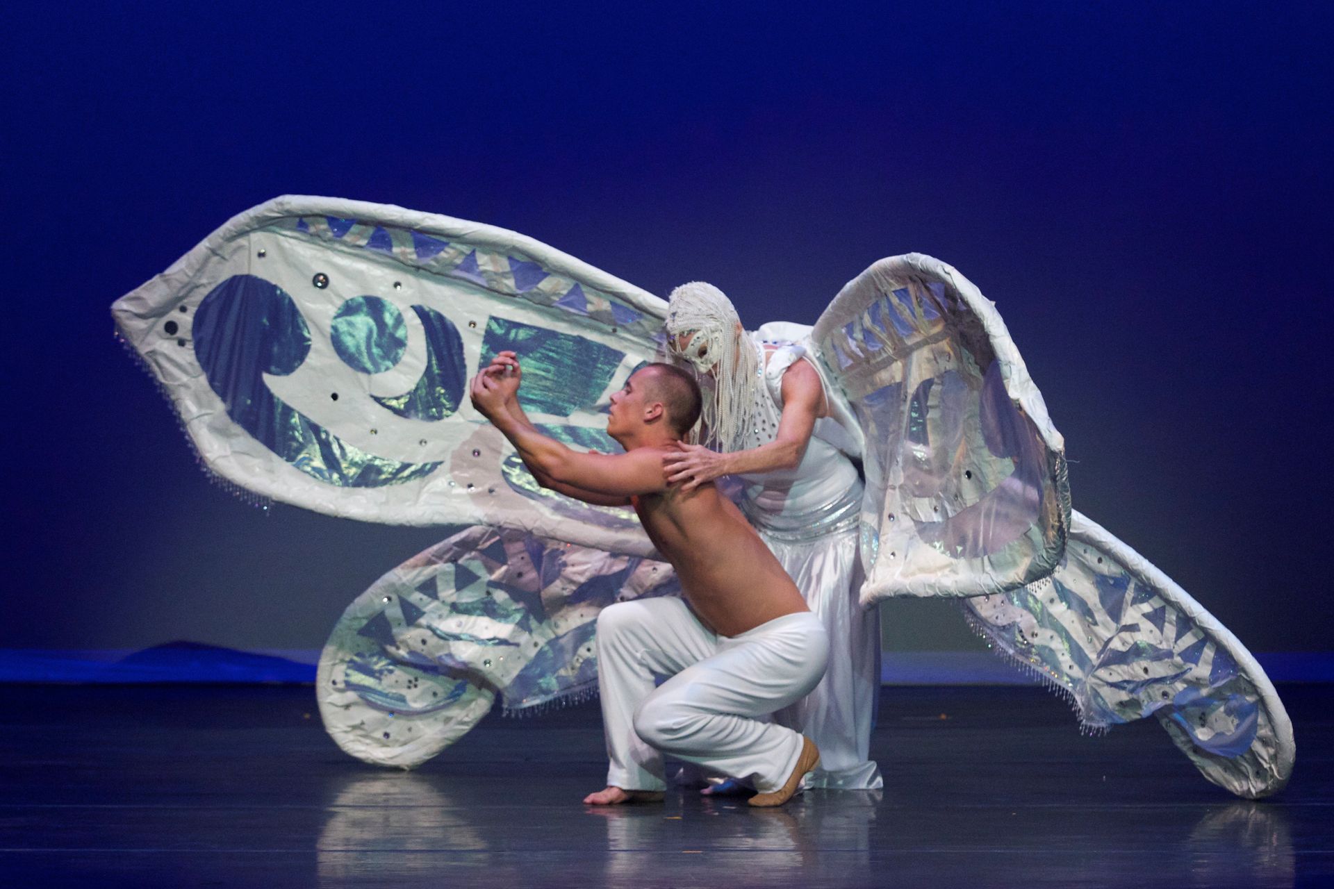 Dancers on stage. One kneels, the other embraces, both under large, white, wing-like fabric. Blue backdrop.