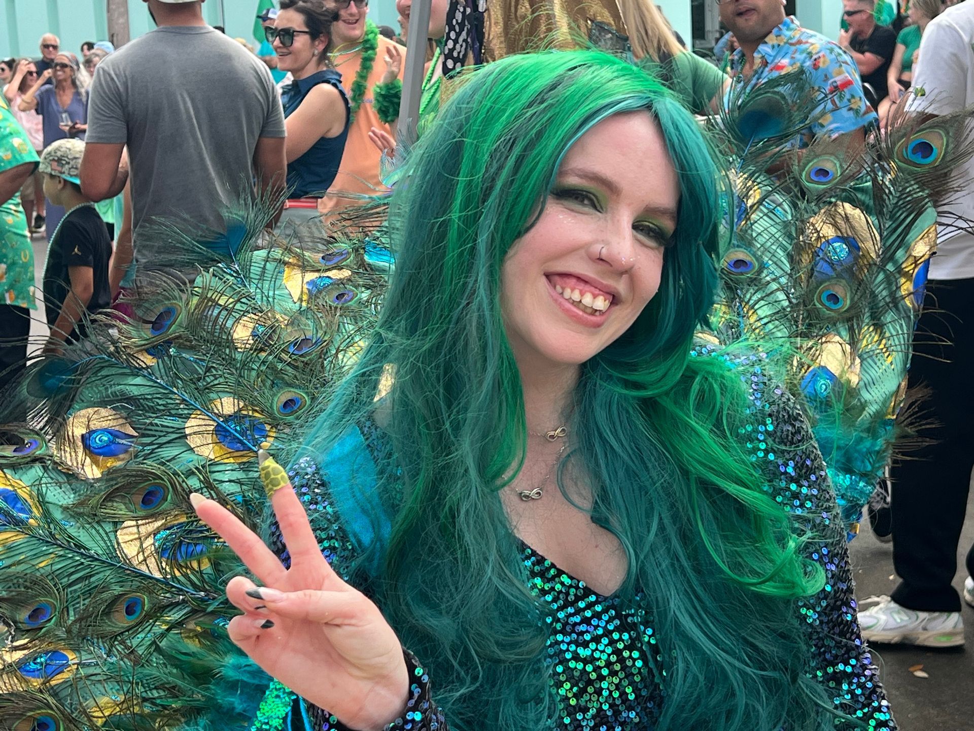 Smiling person in green peacock costume flashing a peace sign at a crowded outdoor event