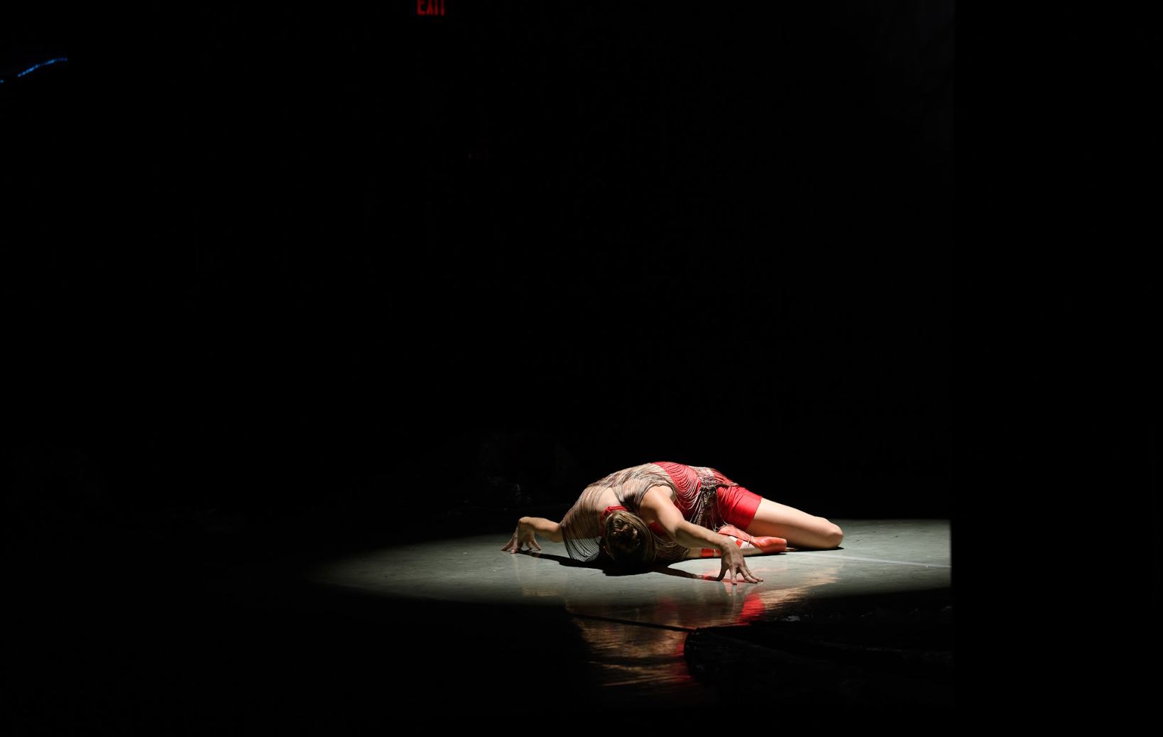 Dancer on stage in spotlight, wearing red; bent on the floor in a pose of distress. Dark background.