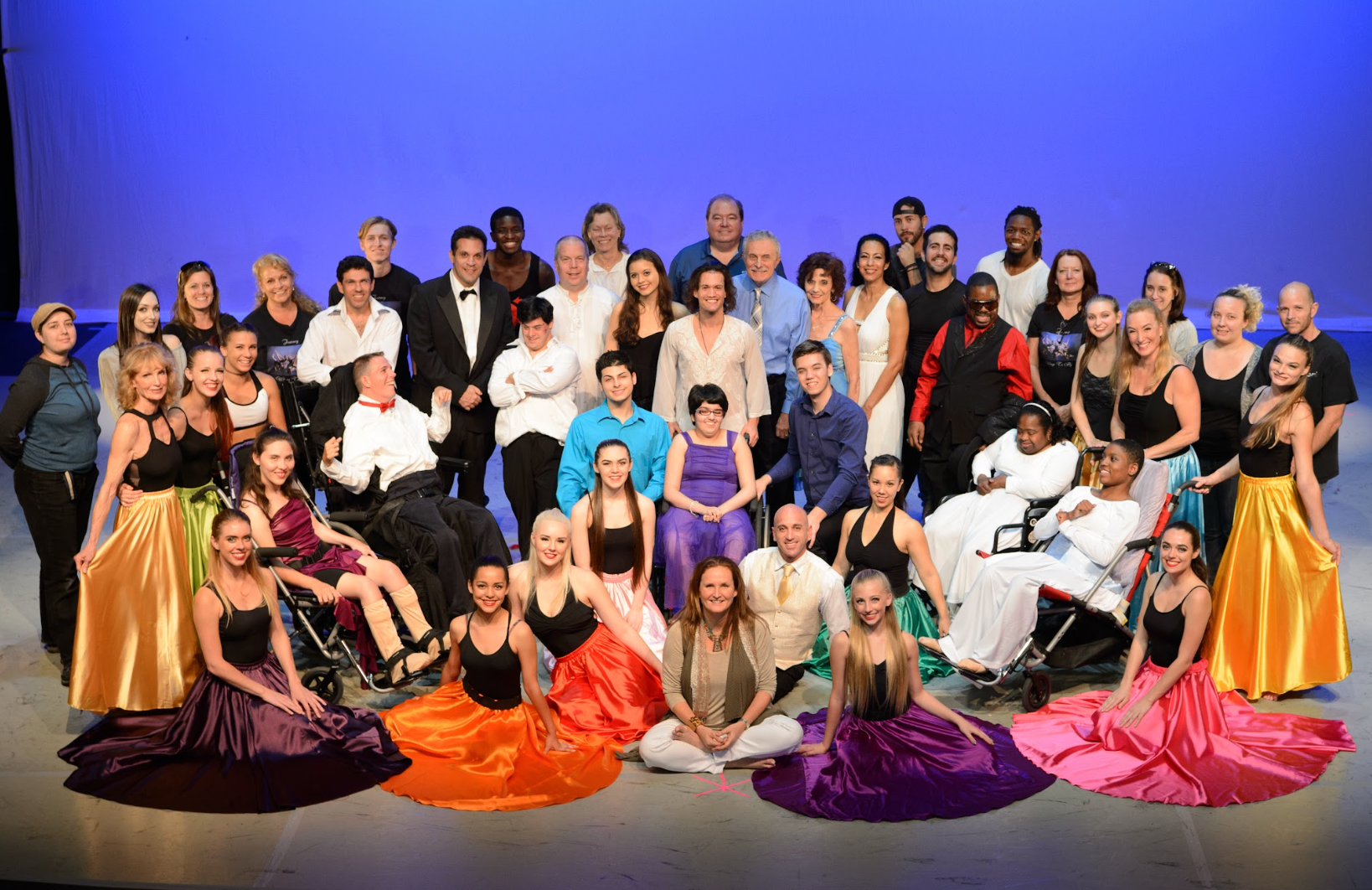 Large group posed on a stage with a blue backdrop, dressed in colorful costumes and formal wear