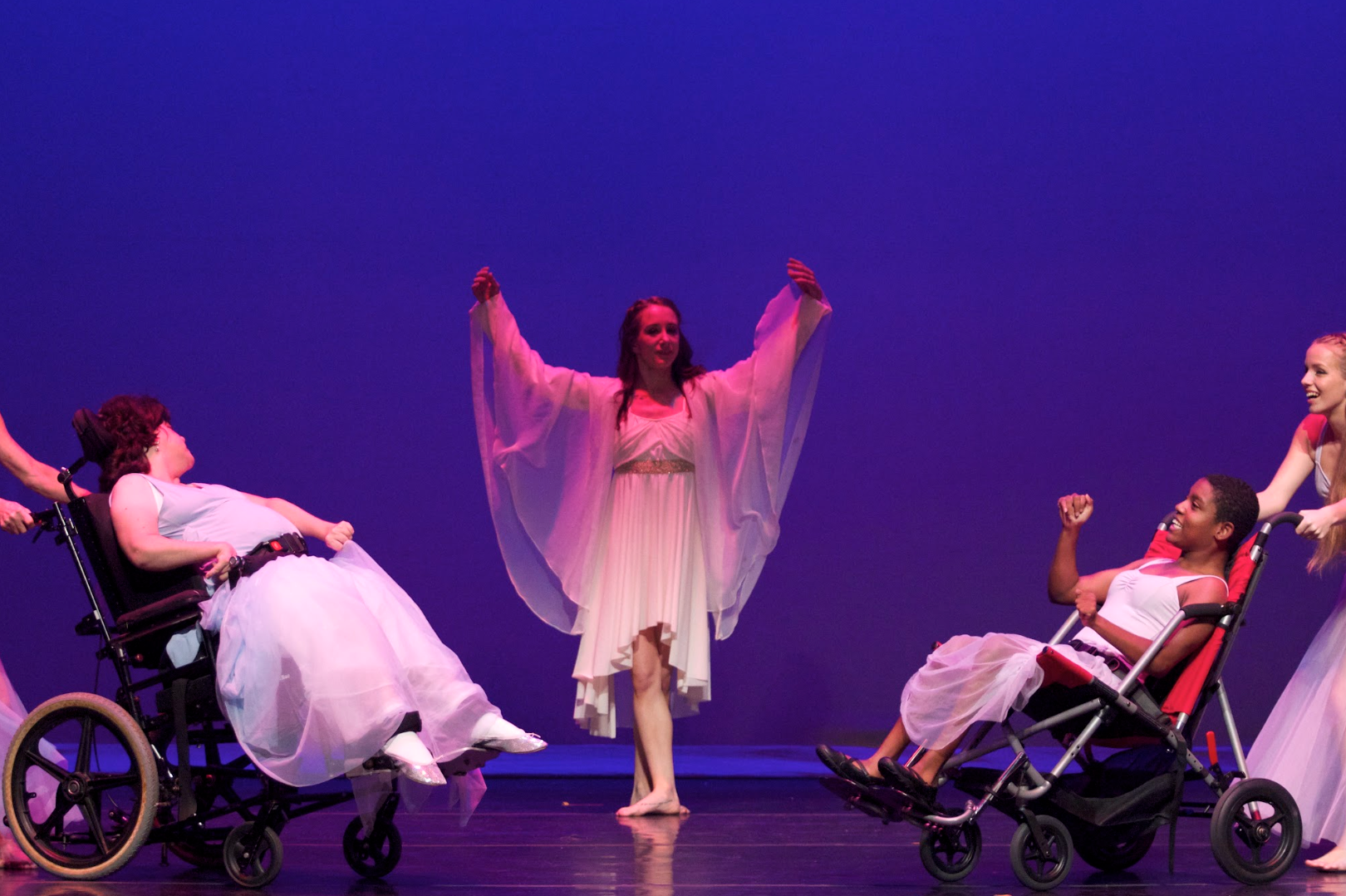 Dancers perform on stage. One in the center with arms up and two in wheelchairs with arms raised.