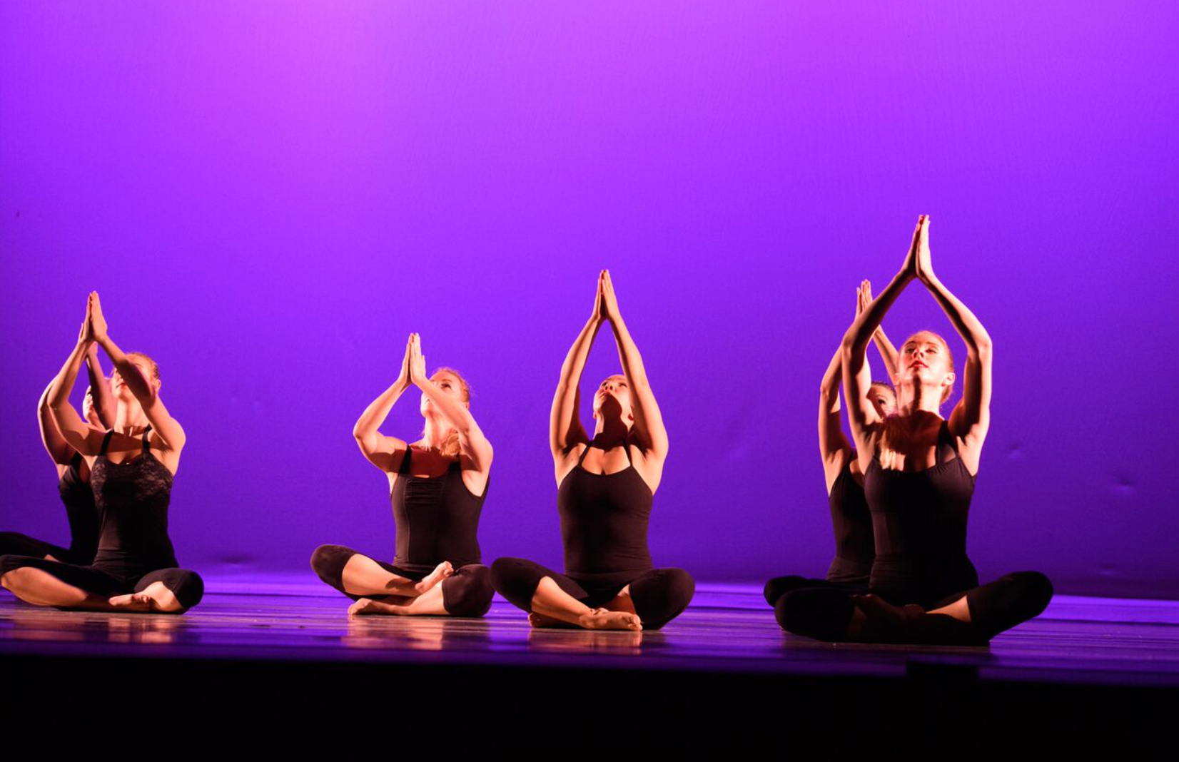 Five people in black, seated in a cross-legged position on stage, hands raised, fingers touching in front of their faces, purple backdrop.