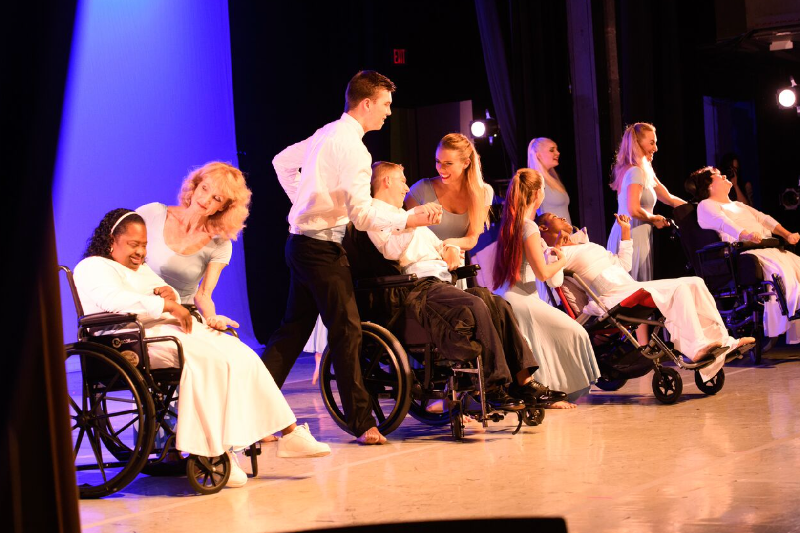 Dancers in wheelchairs and standing performers on stage with blue backdrop, engaging in a dance routine.