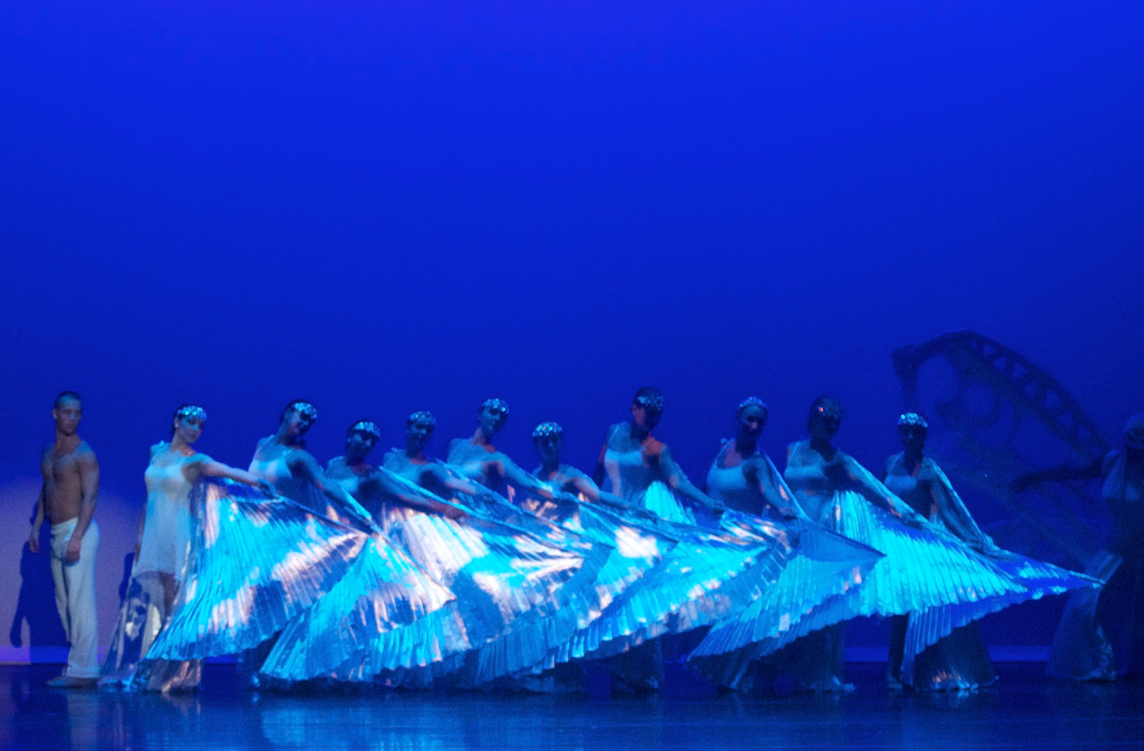 Dancers on stage wearing blue and white costumes, holding large wing-like fabrics, with blue lighting.