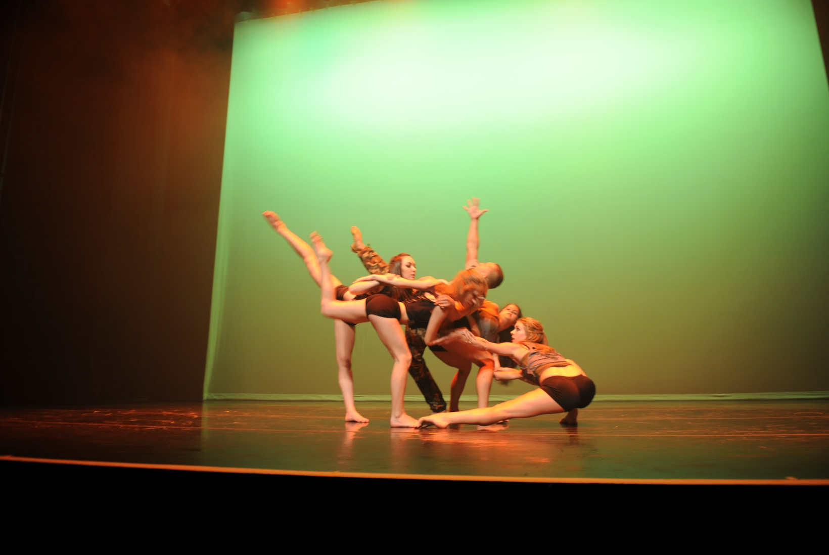 Dancers in brown leotards on a stage with a green backdrop, performing a dynamic dance sequence with various arm and leg extensions.
