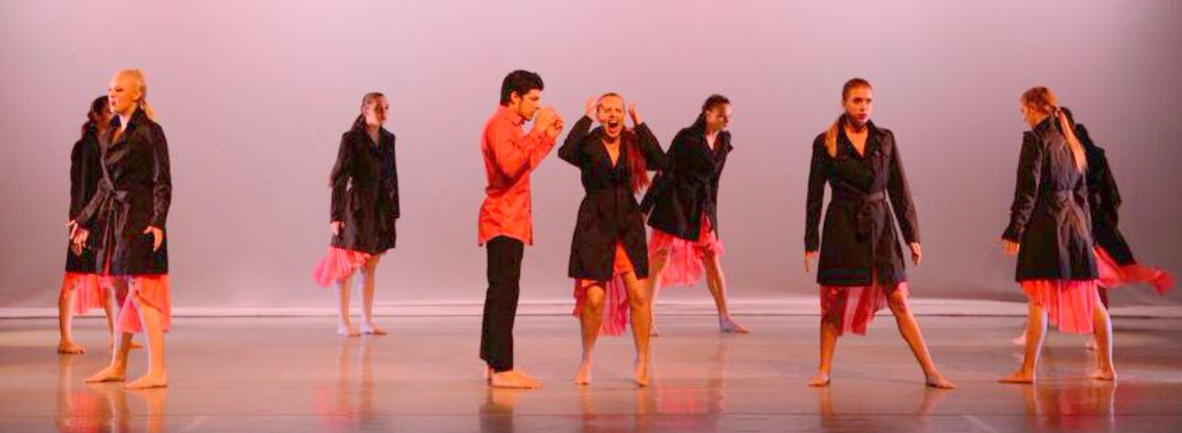 Dancers in dark coats and pink skirts on stage, performing with expressive gestures. One man in an orange shirt.