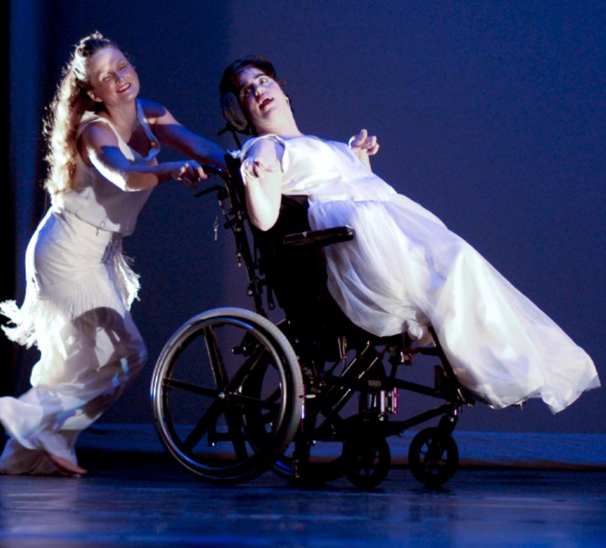 Dancer pushing a person in a wheelchair on stage, both in white dresses, against a blue backdrop.