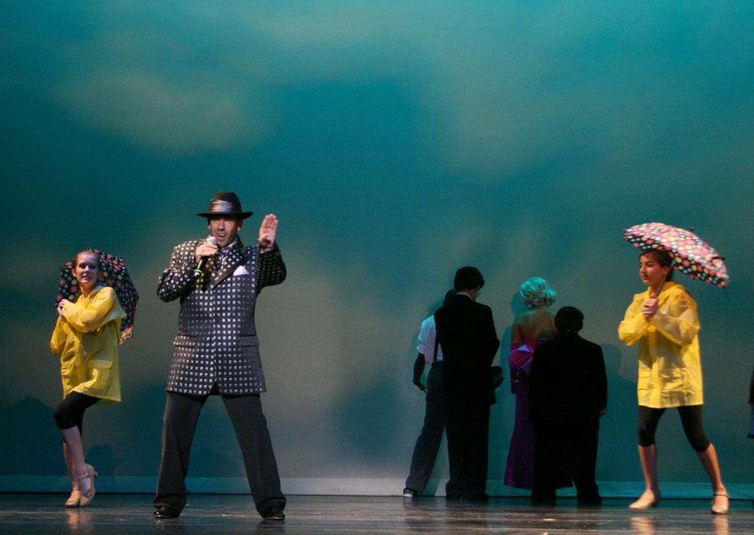 Man singing on stage, wearing hat, stands before dancers in raincoats and umbrellas, blue backdrop.