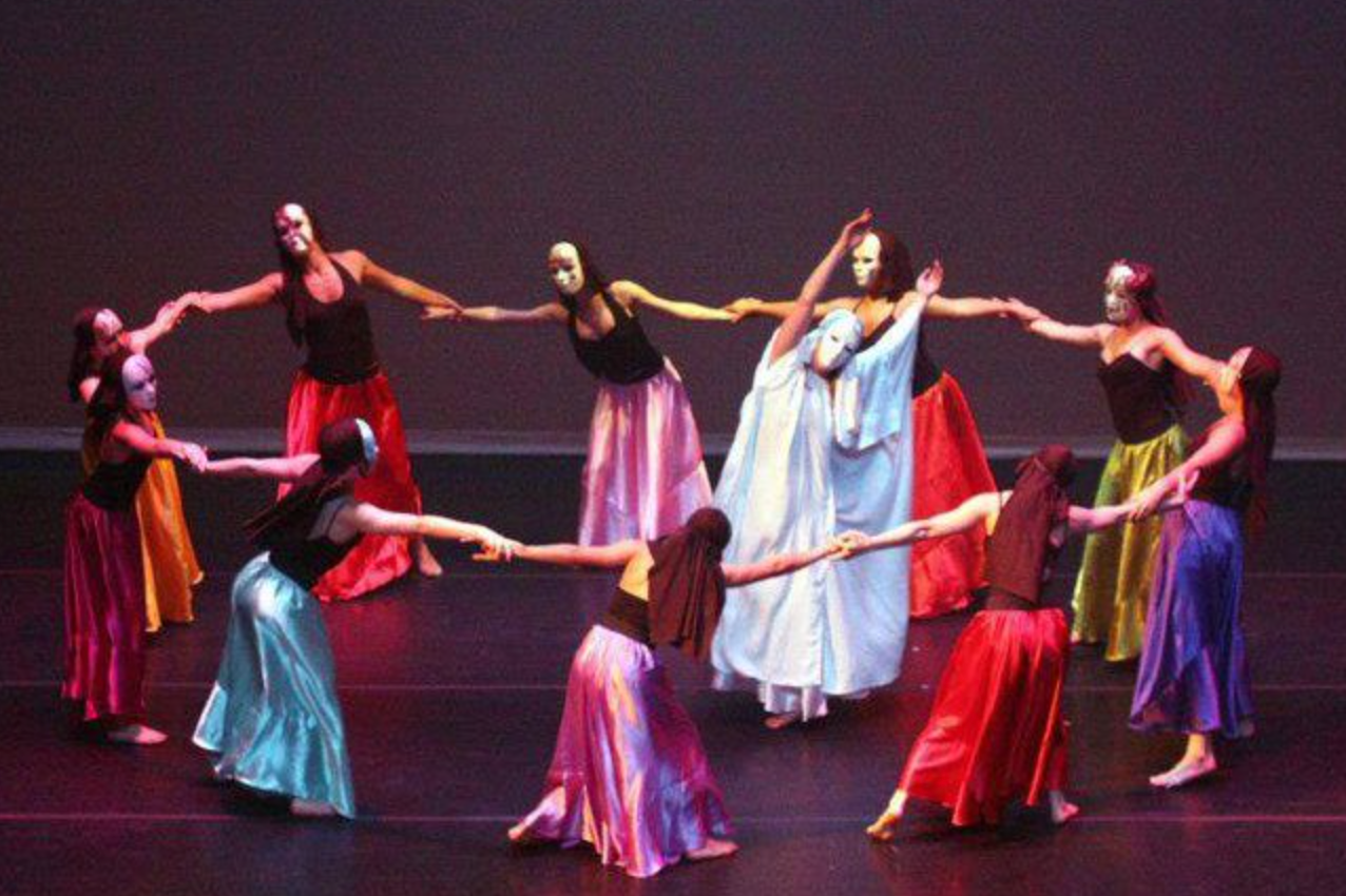 Dancers in colorful skirts and masks form a circle on a stage, holding hands with arms outstretched.