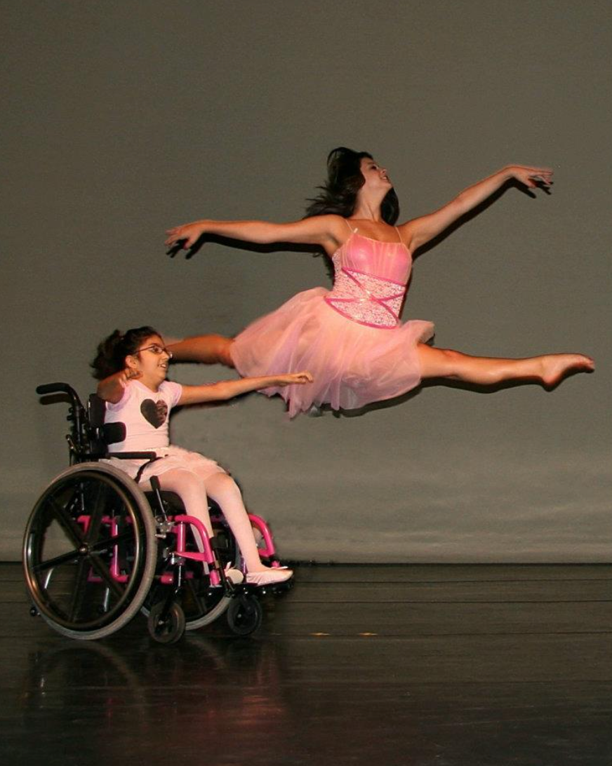 A young dancer in a wheelchair watches a ballerina in a pink tutu jump mid-air on a stage.