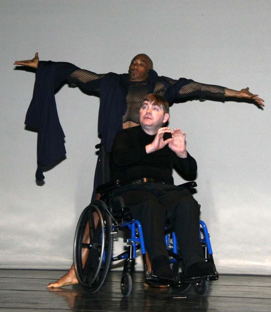 Dancer supports a person in a wheelchair. The standing dancer has arms outstretched, the seated person gestures. Light backdrop.