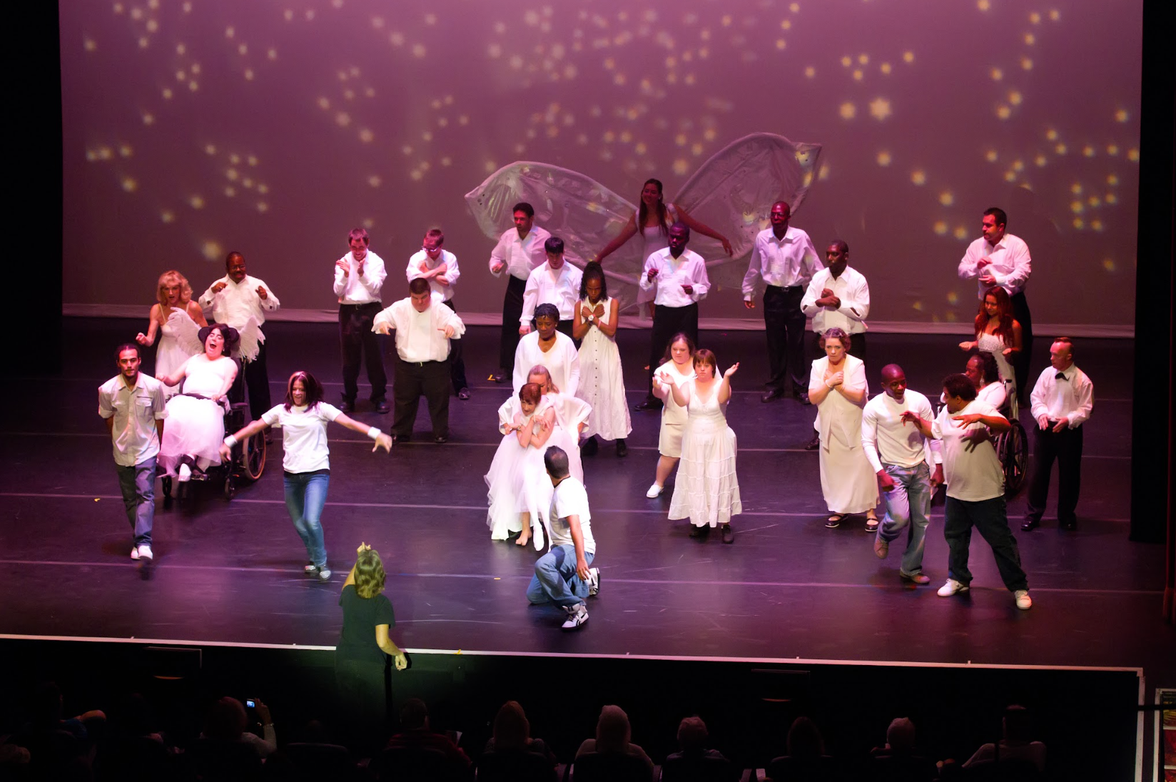 Stage performance with many people in white and black clothing; some dancing, a few with wings.