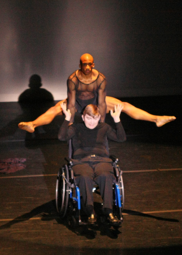 Dancer performs a split over a person in a wheelchair on stage, both in black attire.