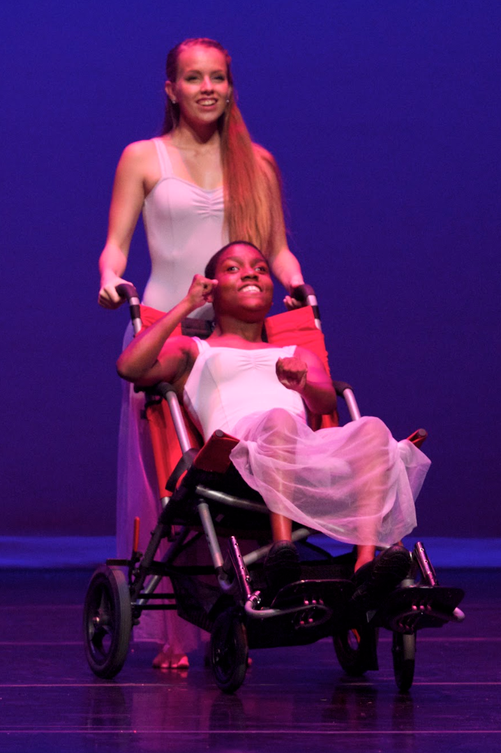 Two dancers on stage, one in a wheelchair, being pushed by another. Both wear white leotards and floaty skirts, against a blue backdrop.