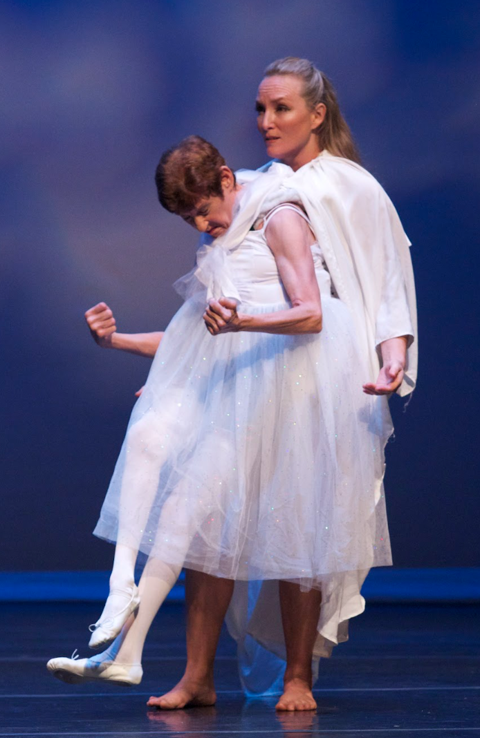 Two people in white costumes performing on a stage with a blue backdrop. One person is supporting the other.