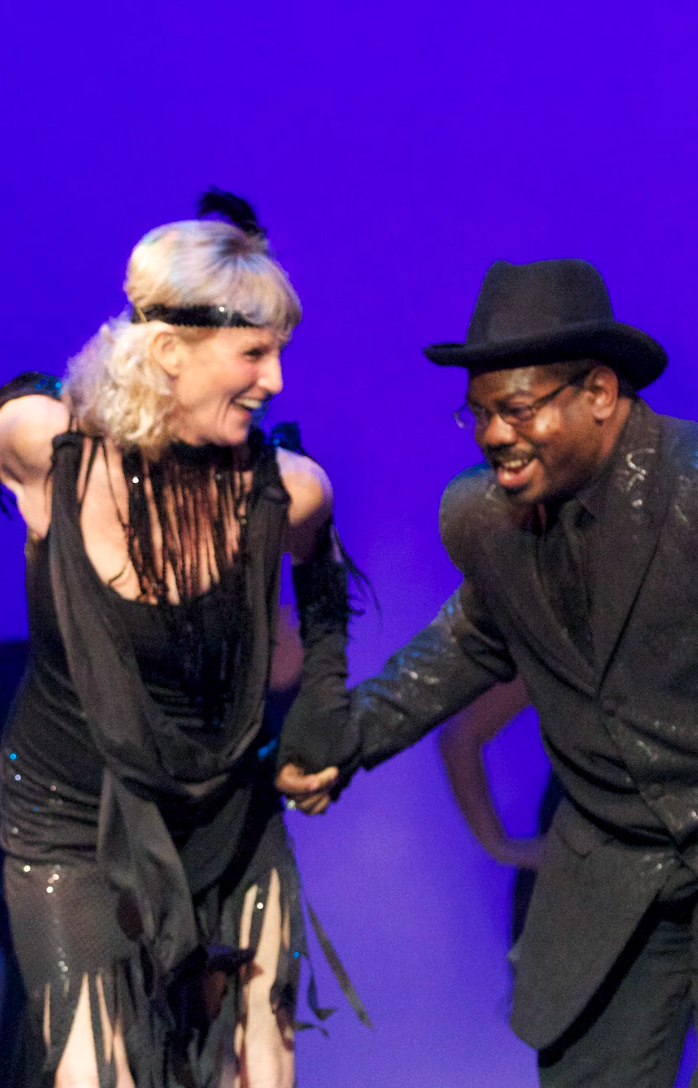 Woman and man dancing on stage, dressed in black, smiling, with a blue background.