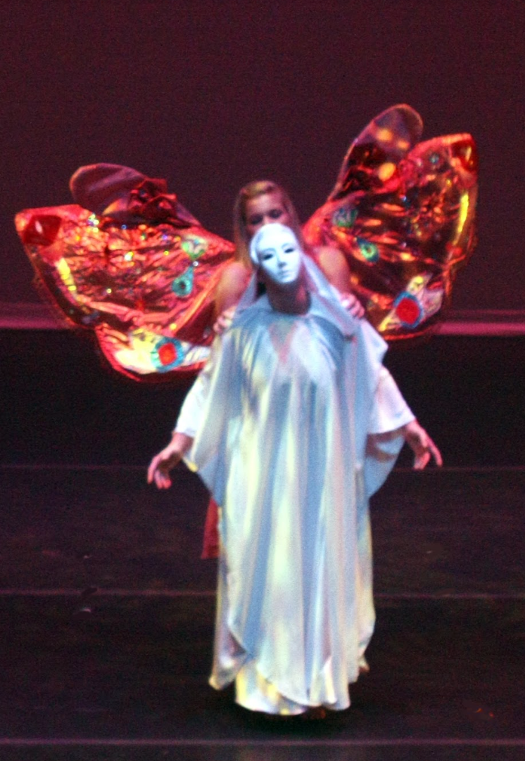 A person in white robes and a mask with butterfly wings in the background on a stage.