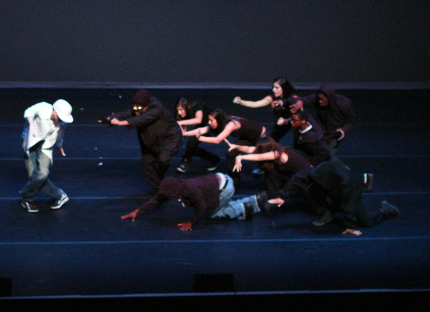 Dancers on stage. One in white jacket, others in dark clothing. All are striking poses, moving toward one.