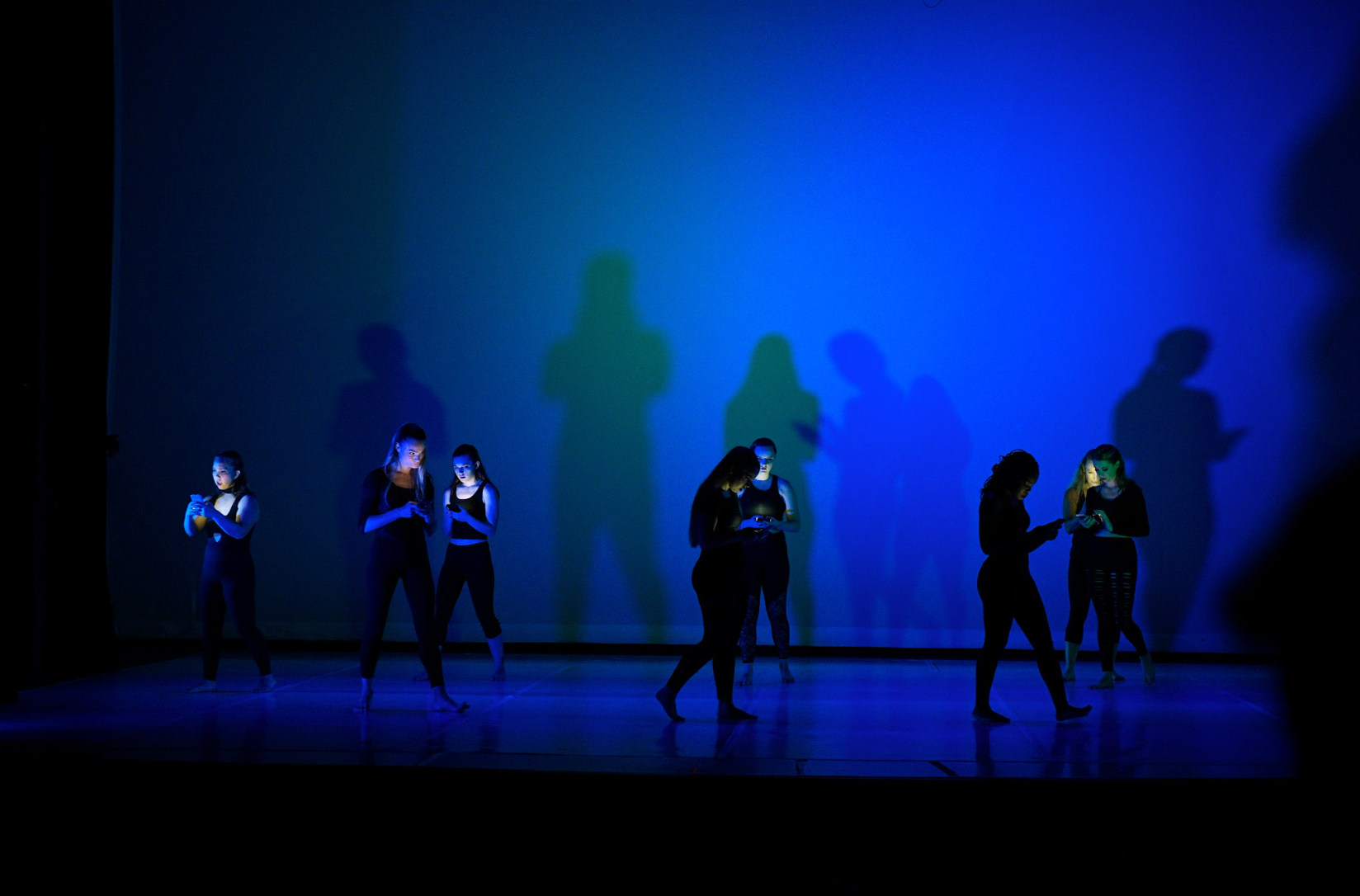 Five dancers on a blue-lit stage with shadowy figures behind them