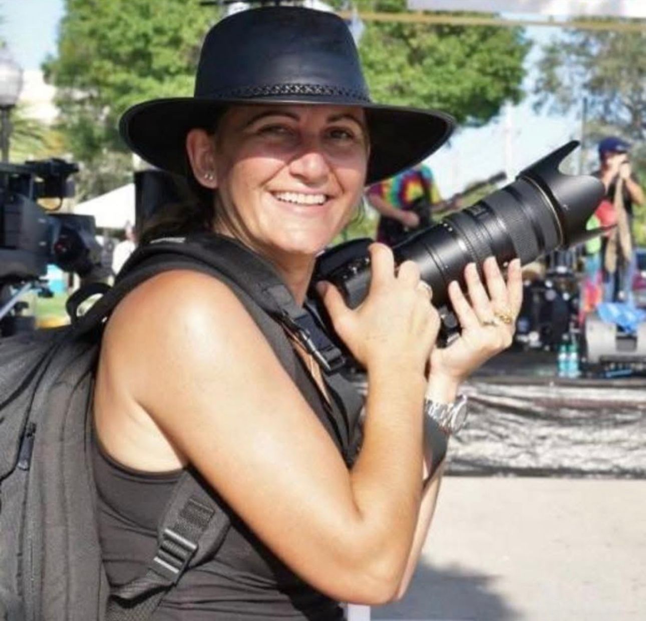 Woman in a black hat holding a large camera with a telephoto lens outdoors, smiling at an event