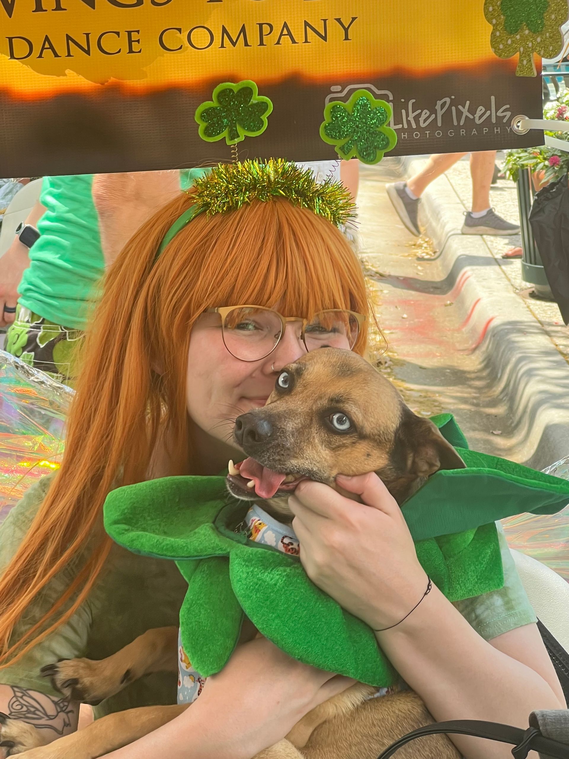 Person in green costume holding a small dog, with orange wig and festive shamrock headband.