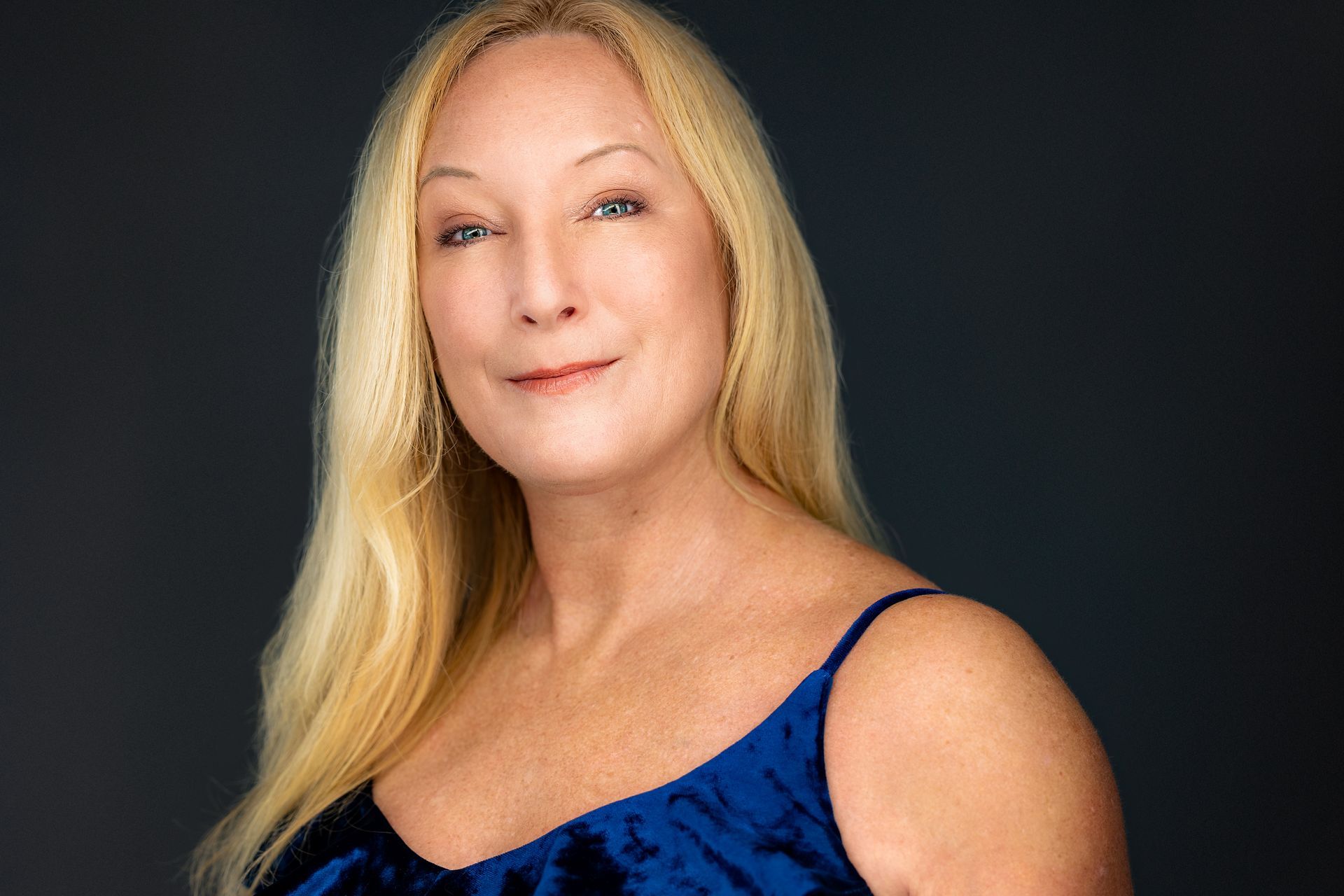Blonde woman with fair skin, wearing a blue velvet top, smiling gently against a dark background.