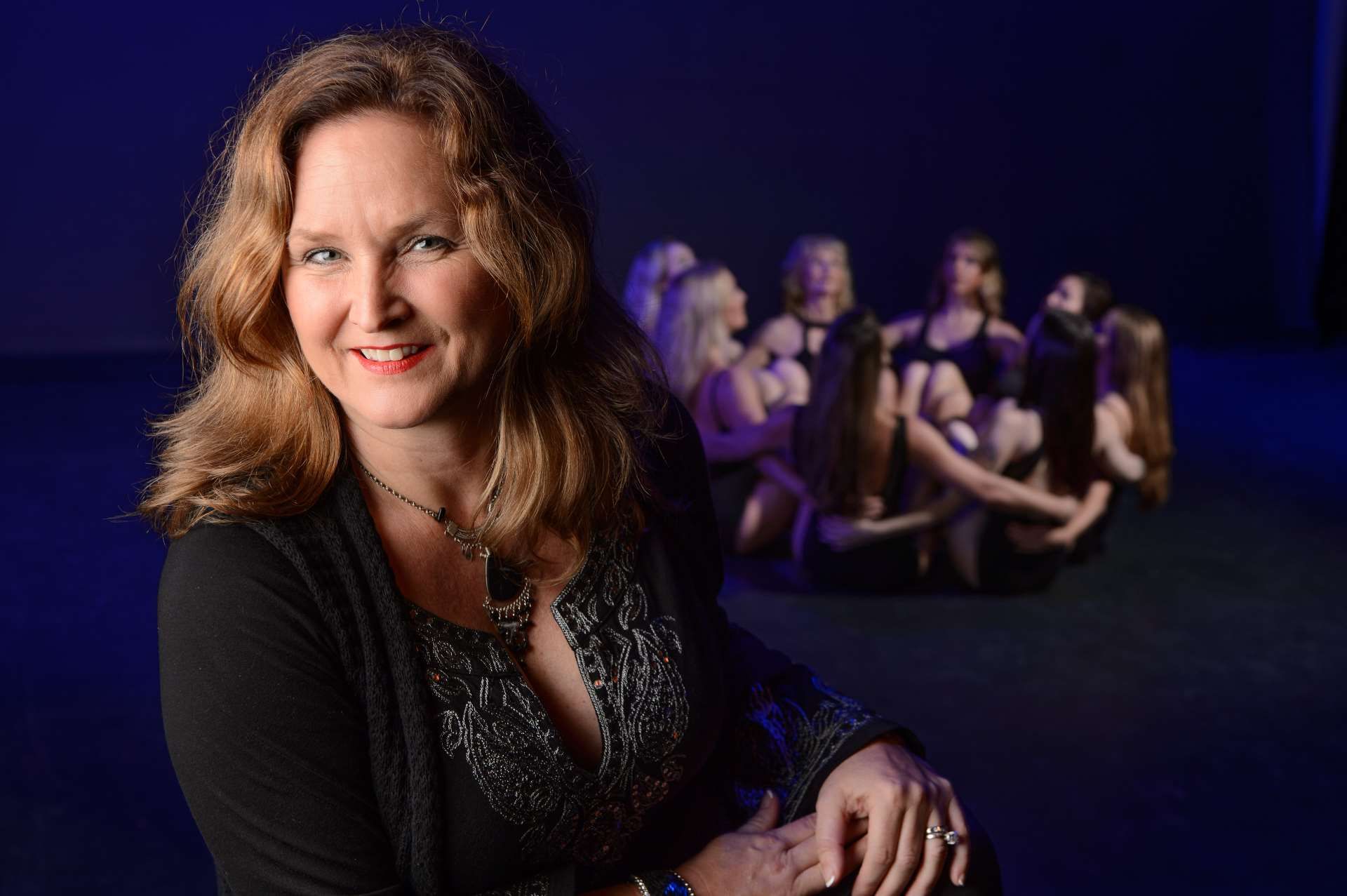 Woman smiles in front of a group of dancers in a blue-lit space.