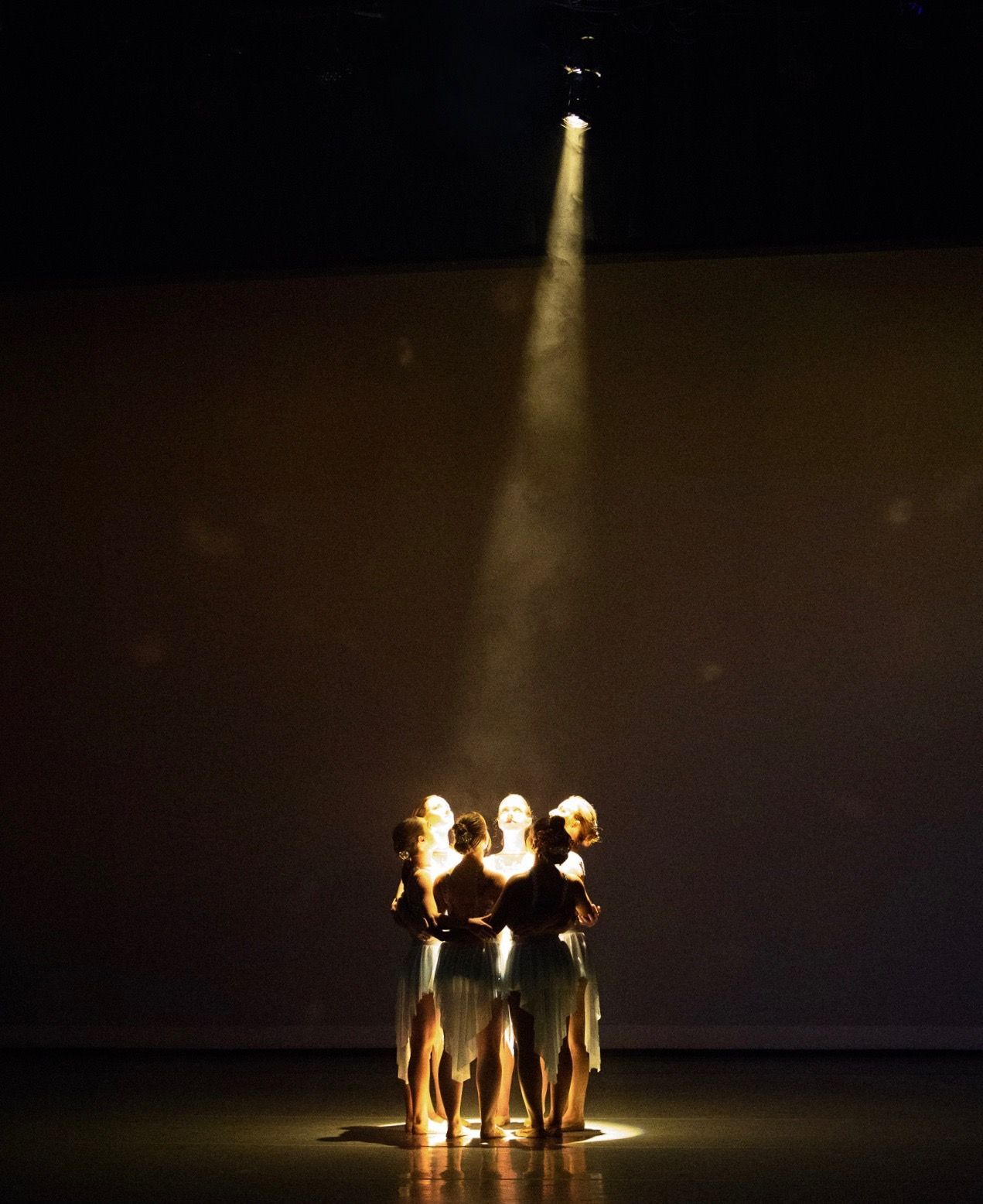 Dancers in a circle, lit by a spotlight. Stage is dark. Light colored costumes.