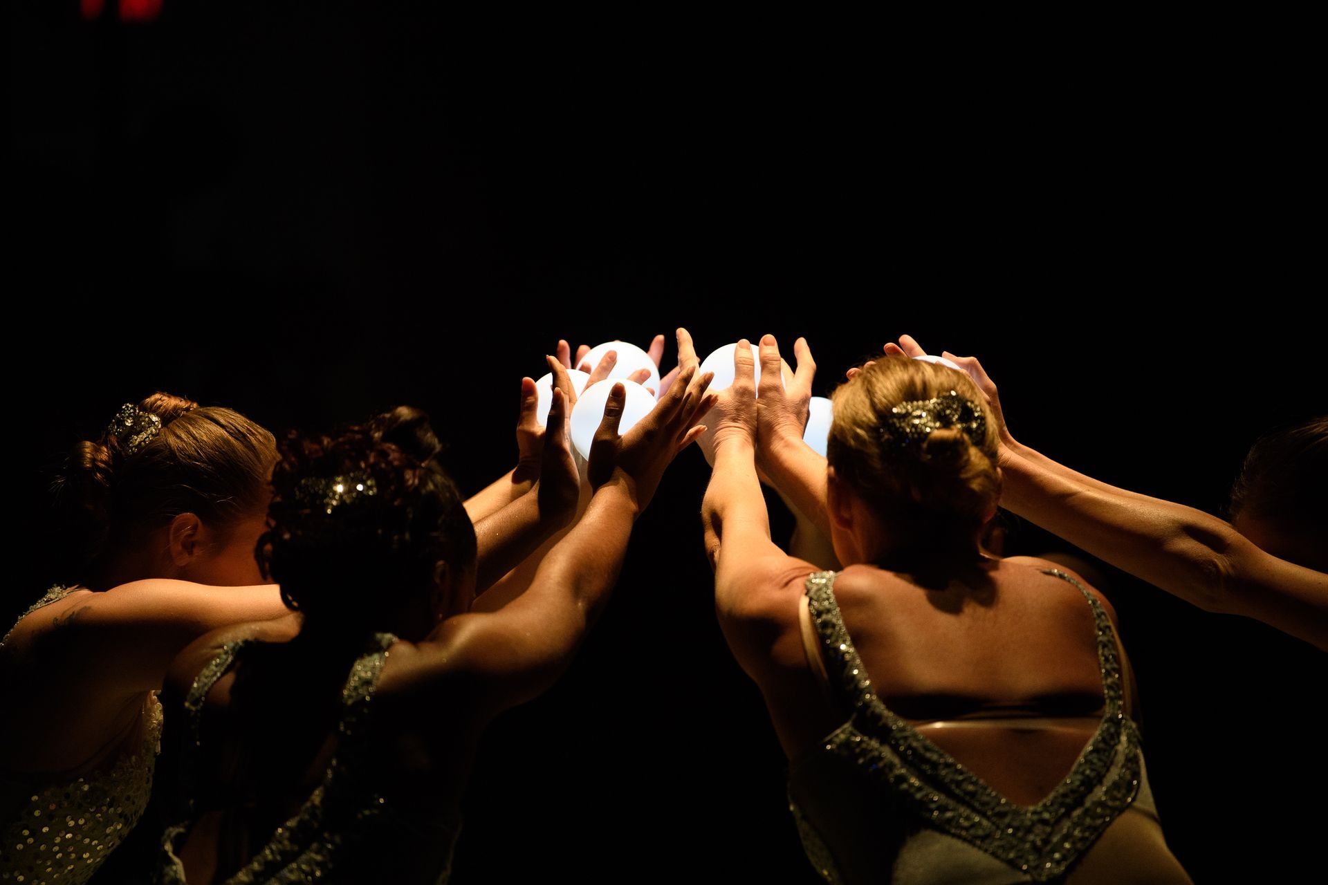 Ballet dancers raise hands around a white object in a dimly lit setting.