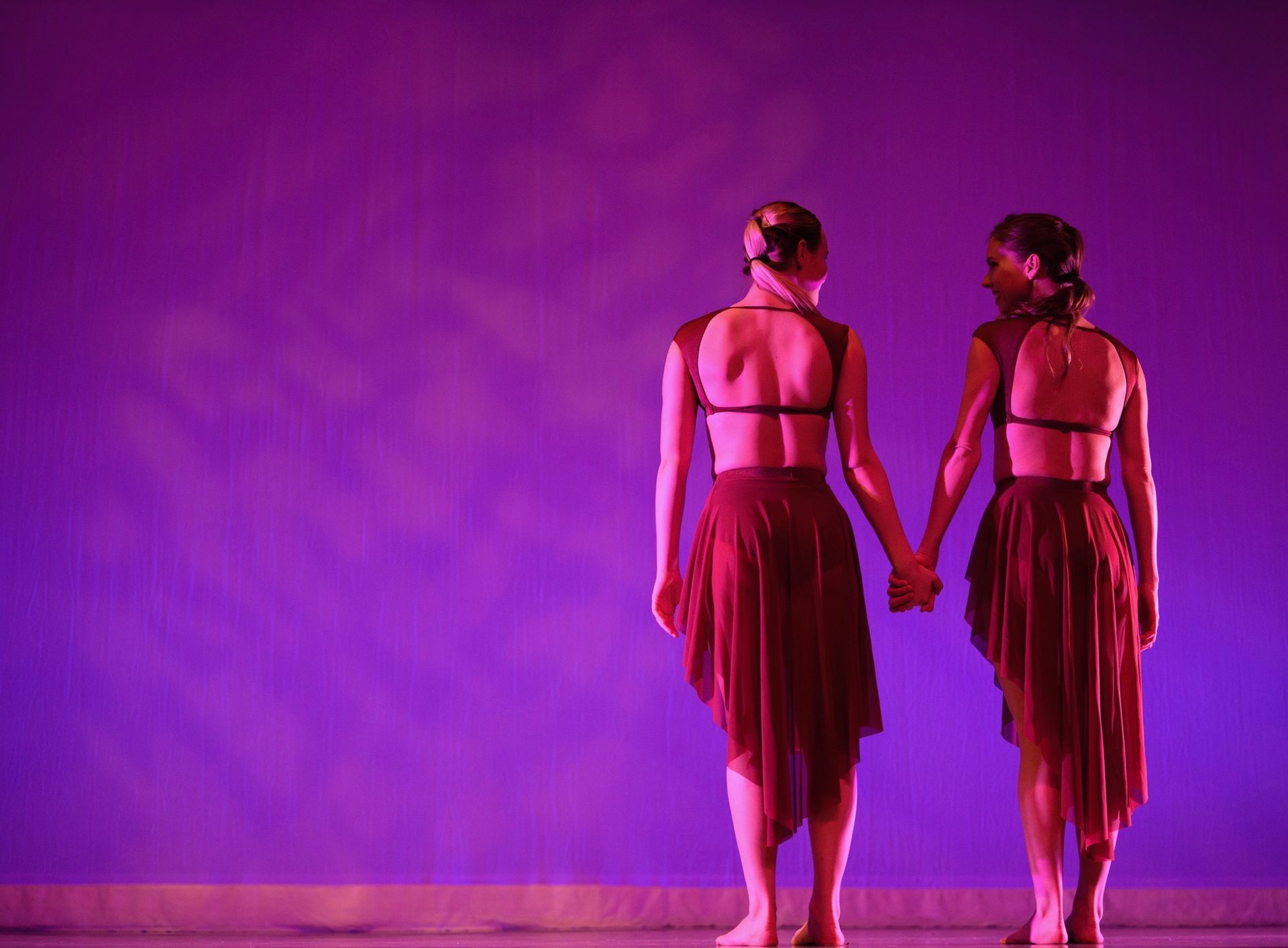 Two dancers in maroon dresses hold hands on a purple stage, backs to the viewer.