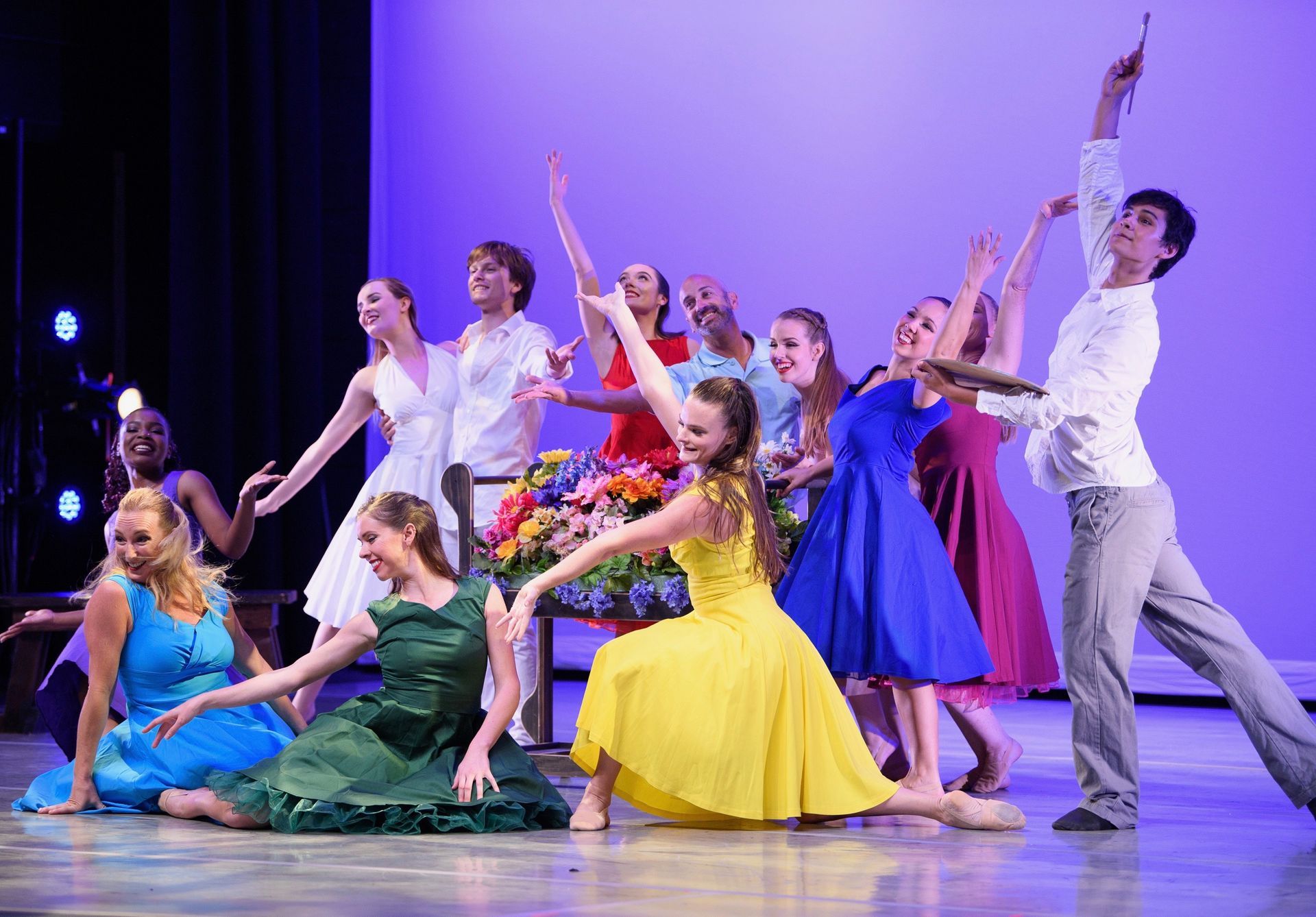 Dancers on stage with arms raised. Bright dresses, flowers, and a spotlight on a purple background.