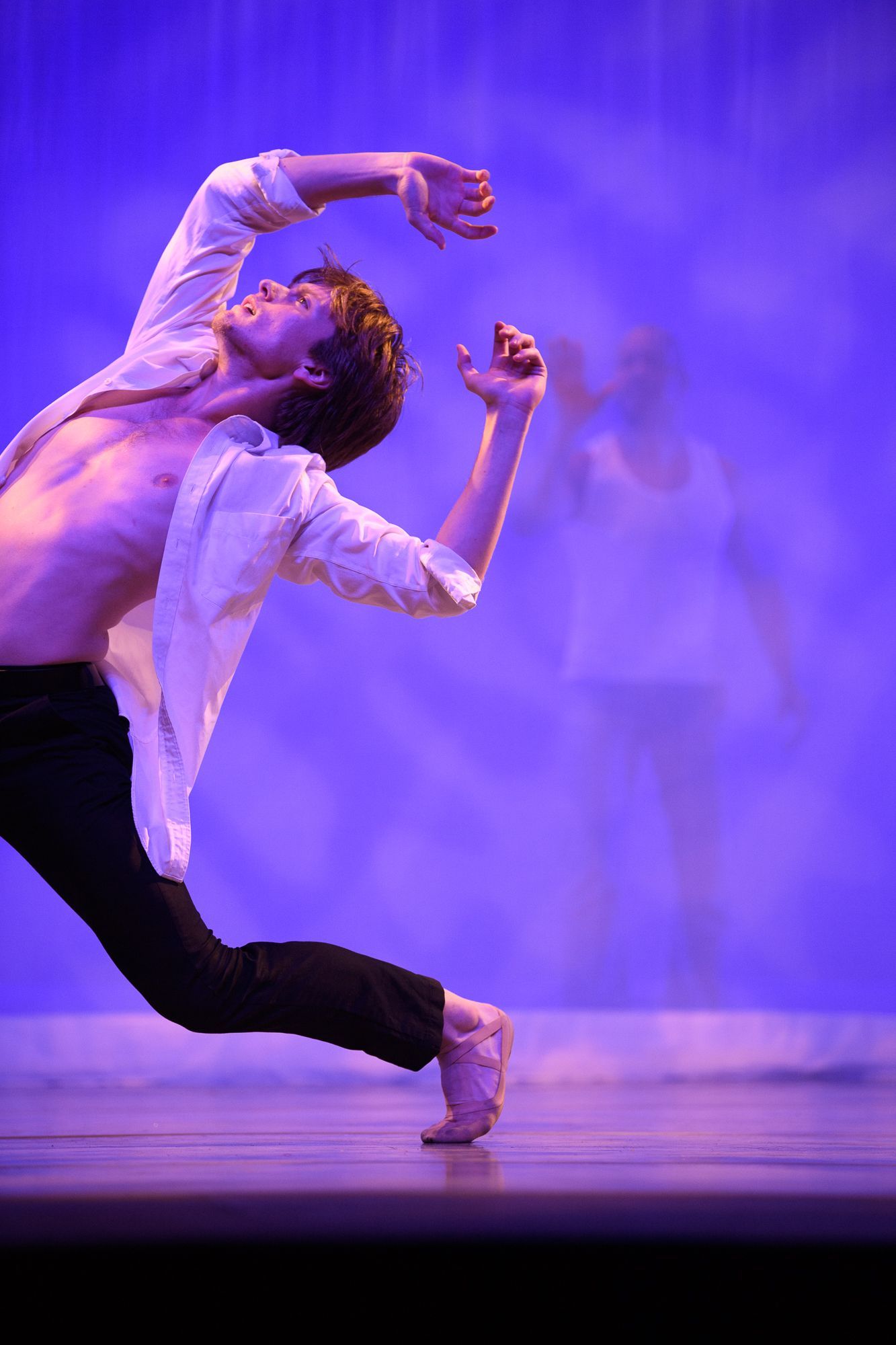 Male dancer in a white shirt and black pants, arched back, arms raised, performing on stage with blue lighting.