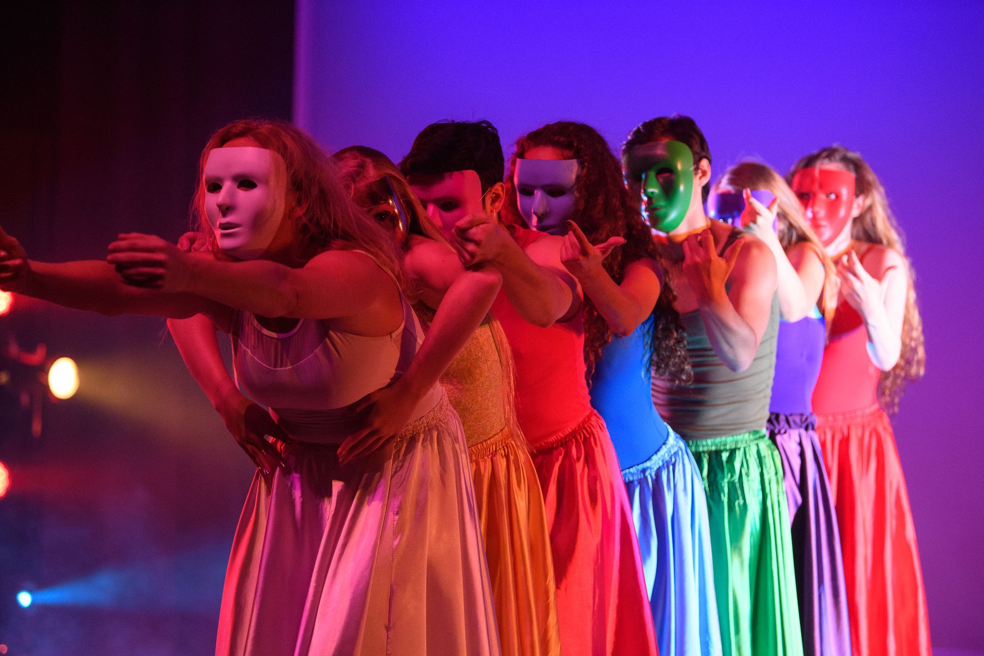 Dancers in colorful gowns and face paint, wearing masks, performing onstage under colored lights.