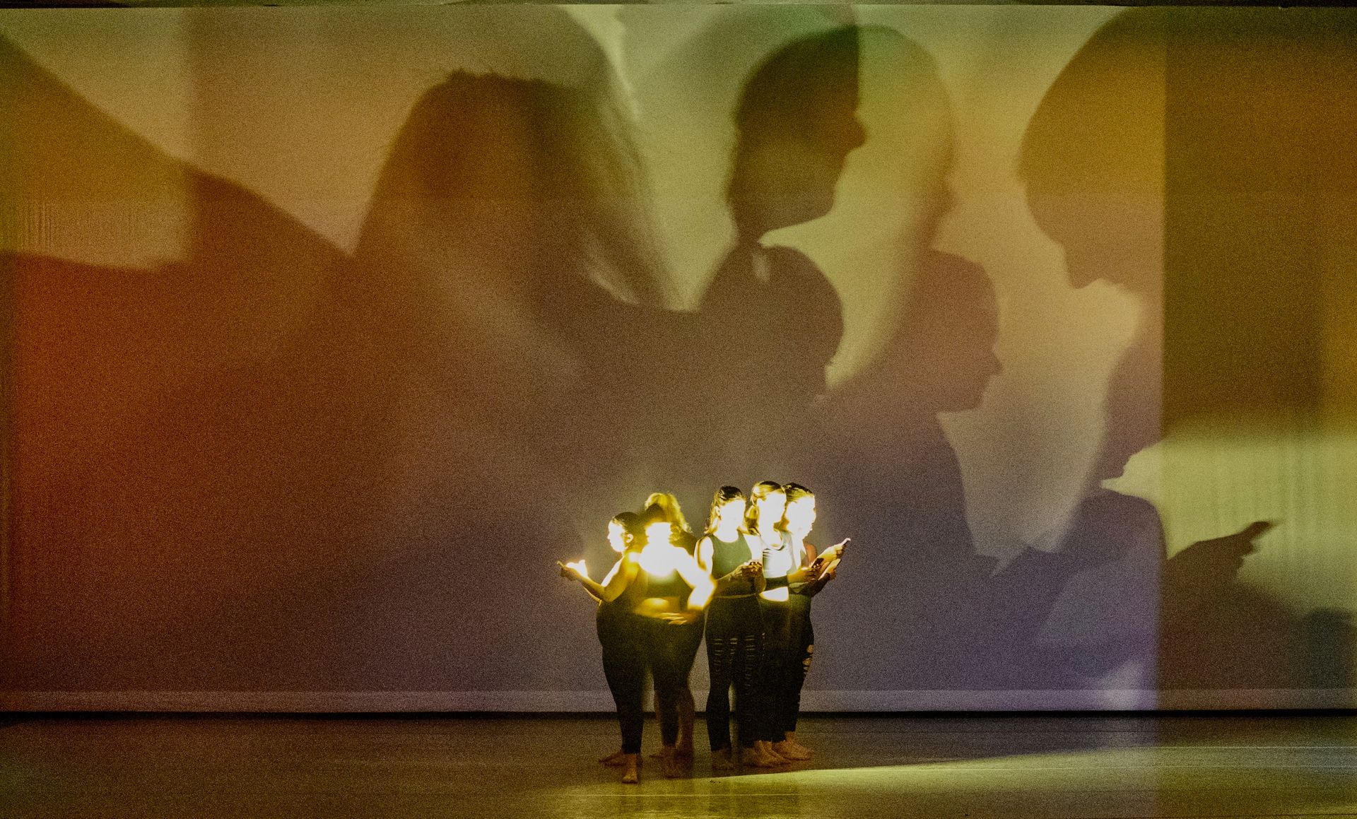 Dancers on stage with colorful shadows projected behind them. Lighting creates a warm glow.