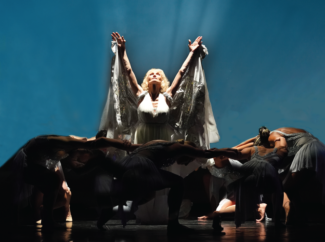 Dancers in black costumes kneel around a central performer raising her arms against a blue background
