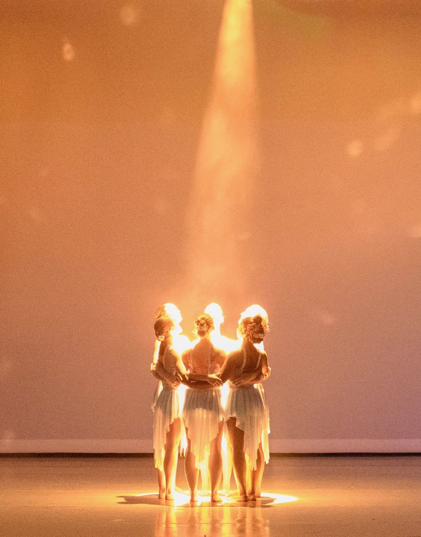 Three dancers on stage, backlit by a spotlight. They hold hands, wearing white costumes, illuminated in golden light.