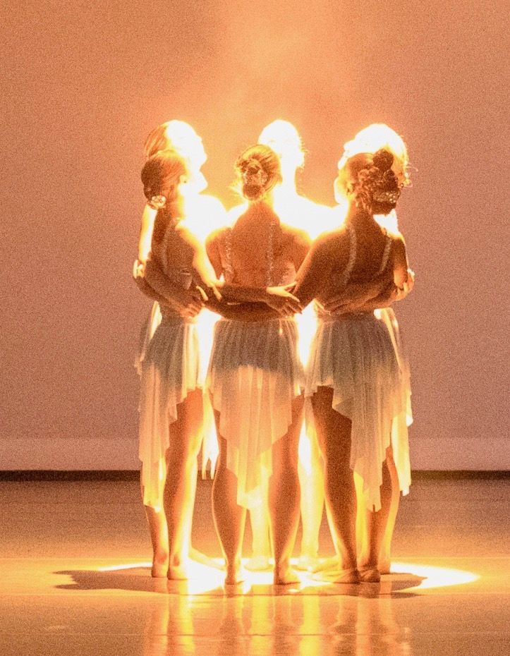 Dancers in white dresses huddle in a circle, bathed in warm light on a stage.