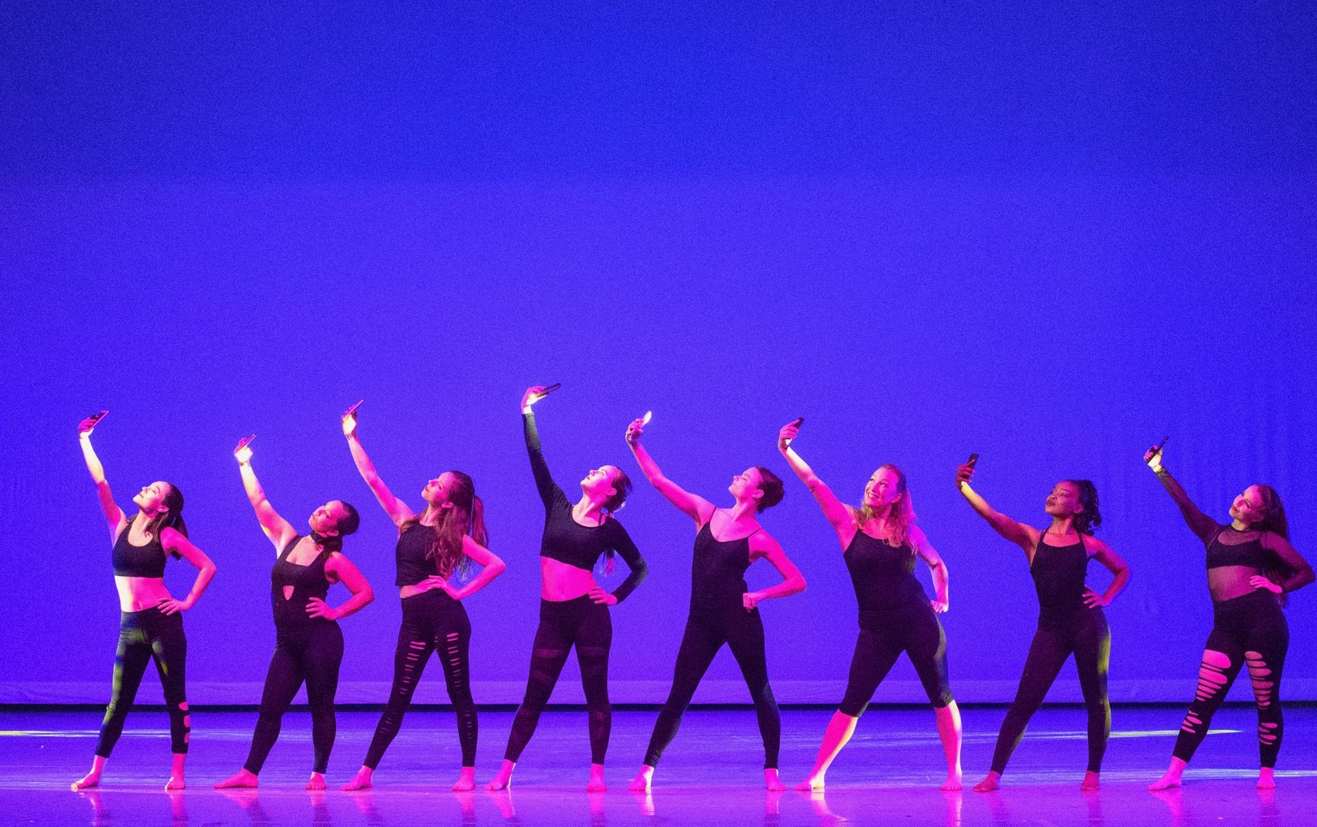 Dancers on stage with arms raised. They wear black outfits against a blue backdrop.