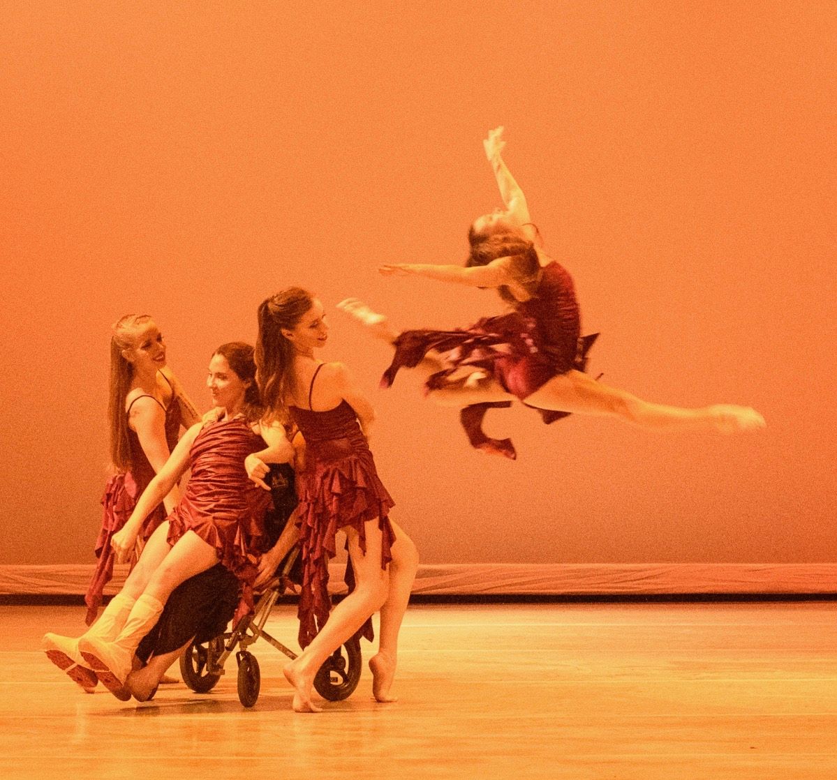 Dancers in maroon costumes on stage; one in wheelchair, others supporting; one dancer leaps. Orange background.