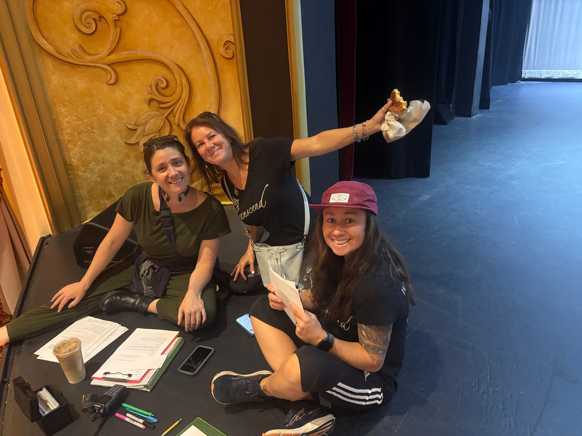 Three people sitting on a stage floor, smiling and posing with papers and a phone near a curtain backdrop